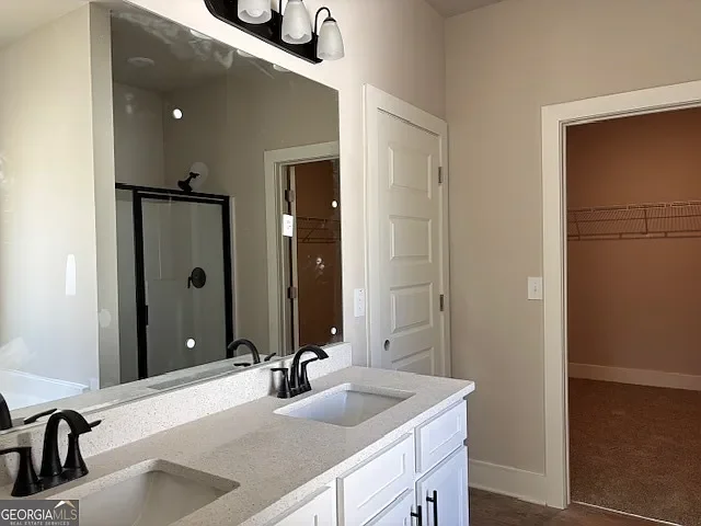 Bathroom with dual sinks, large mirror, white cabinets, light fixtures, door to a closet or storage area, and a walk-in shower with glass door.