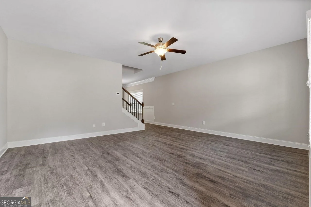 Empty living room with gray walls, wood flooring, ceiling fan, and staircase.