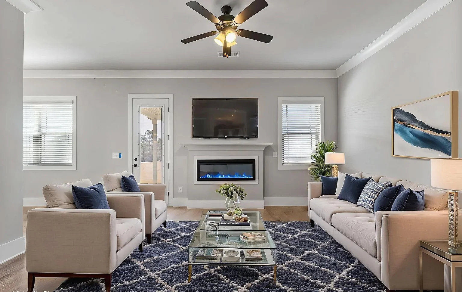 Living room with two beige sofas decorated with navy blue pillows, a glass coffee table, a fireplace, a wall-mounted TV, windows with blinds, a ceiling fan, a door leading outside, and a framed landscape painting.