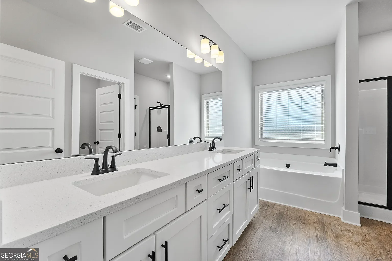 Modern bathroom with white cabinetry, double sinks, black fixtures, a large mirror, a bathtub, a window with blinds, and wood-look flooring.