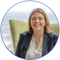 A woman with shoulder-length hair smiling, sitting in a green chair, wearing a blazer and white shirt, indoors with a light-colored background.