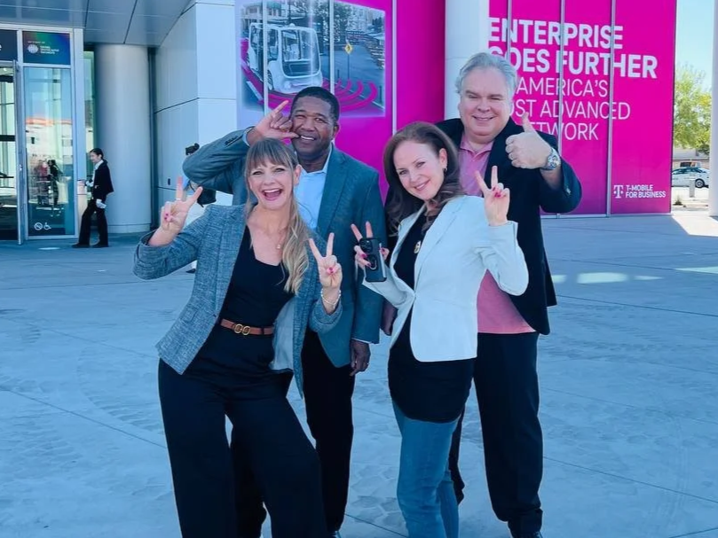 Group of four diverse people smiling and making peace signs outside a T-Mobile store, with a pink advertisement in the background.