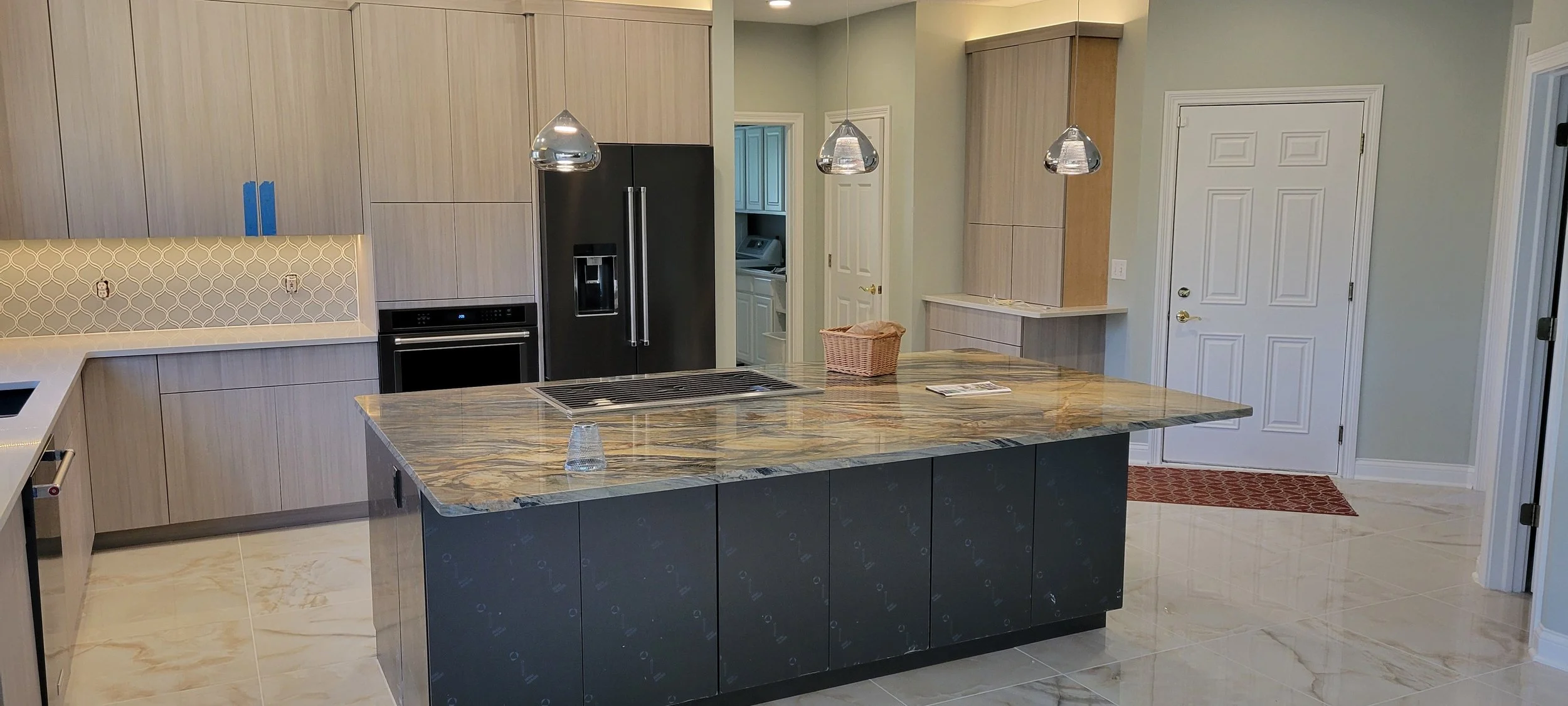 Modern kitchen with marble island counter, black refrigerator, built-in oven, beige cabinets, pendant lighting, laundry area in background, and a white door with a red patterned mat.