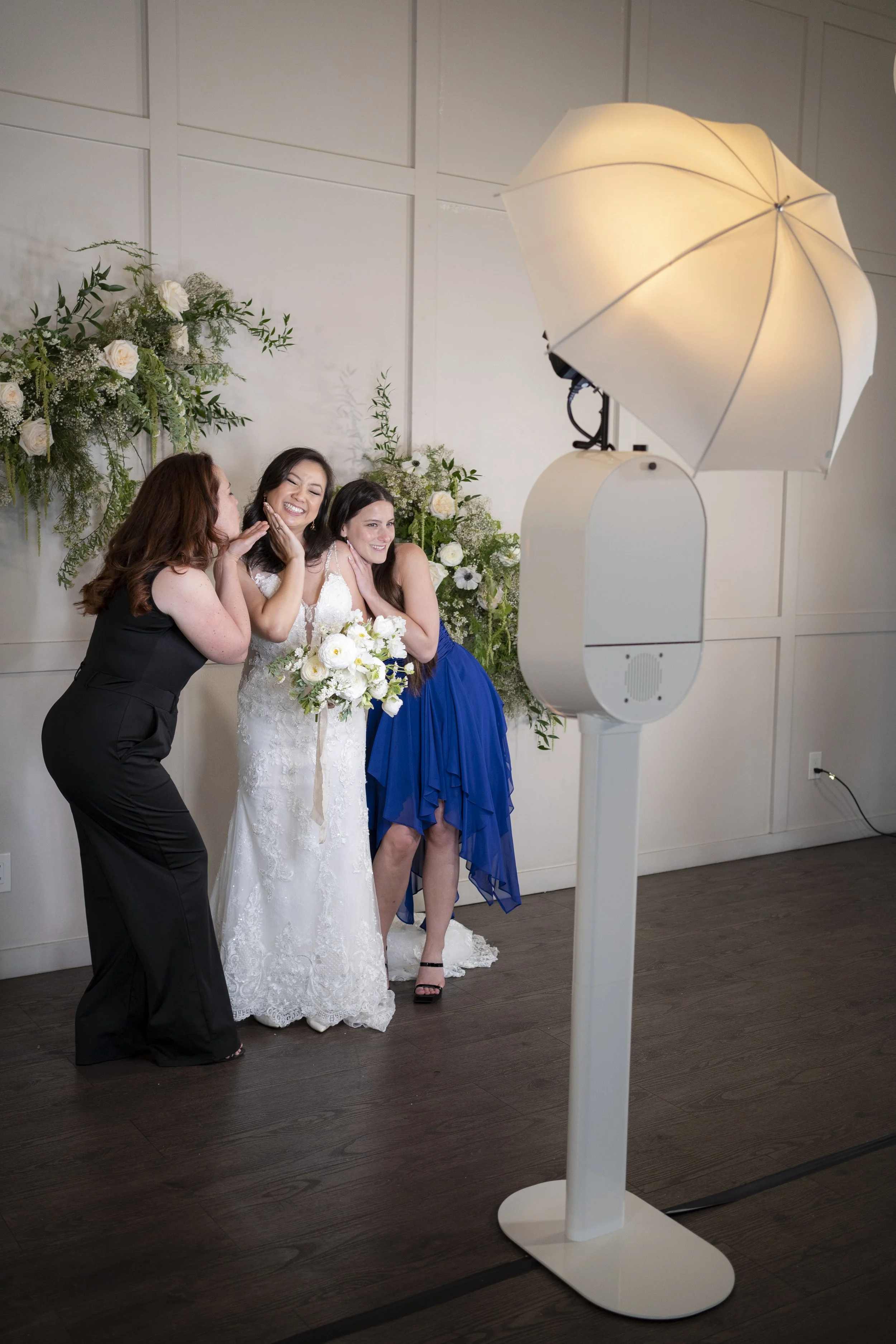 Three women, including a bride in a white wedding dress holding a bouquet, taking a photo with a photo booth using a large professional camera with a white umbrella light. They are smiling and posing happily in front of a floral backdrop with white flowers and greenery.