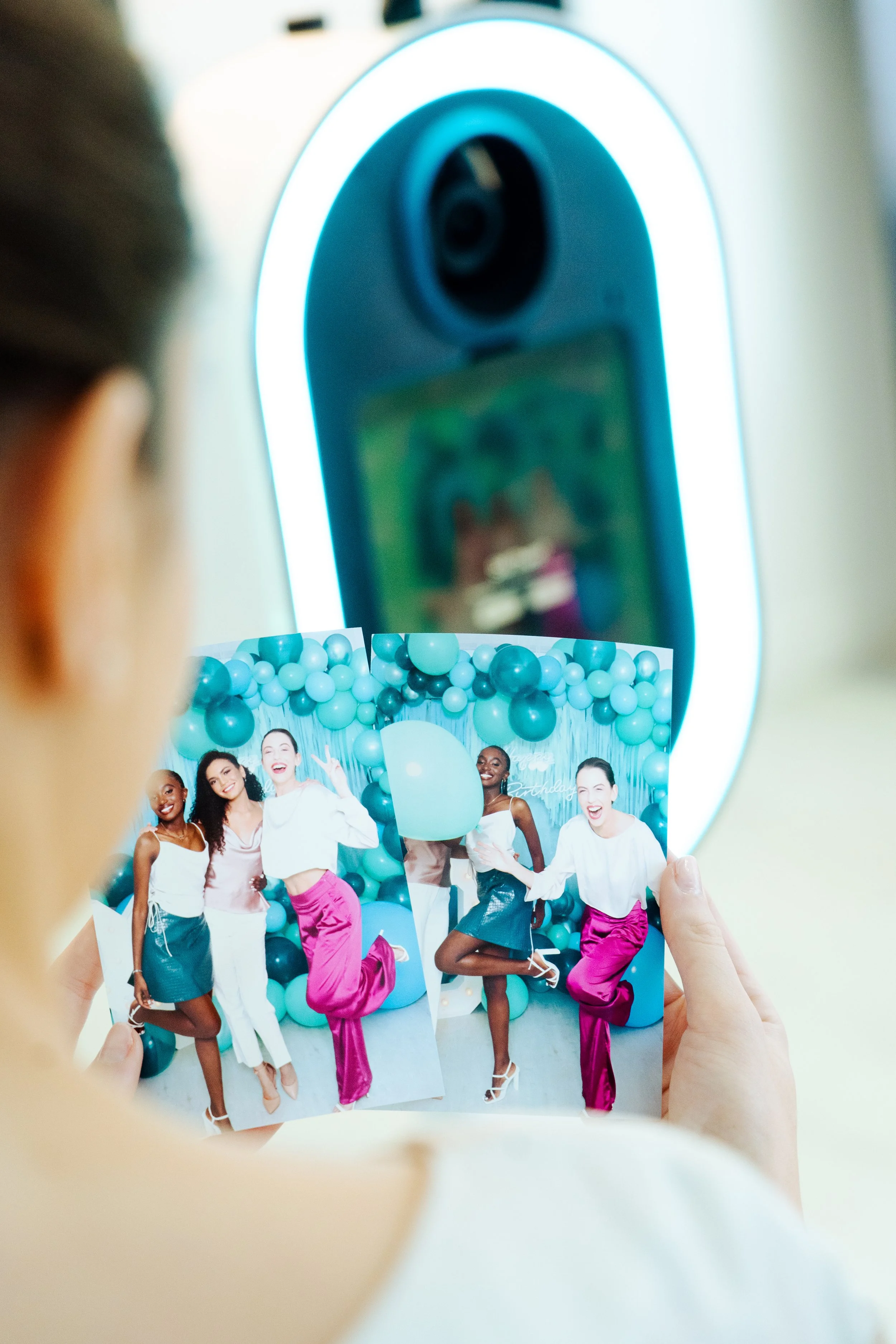 Person holding up two printed photos of a group of women celebrating a birthday, with a balloon backdrop behind them.