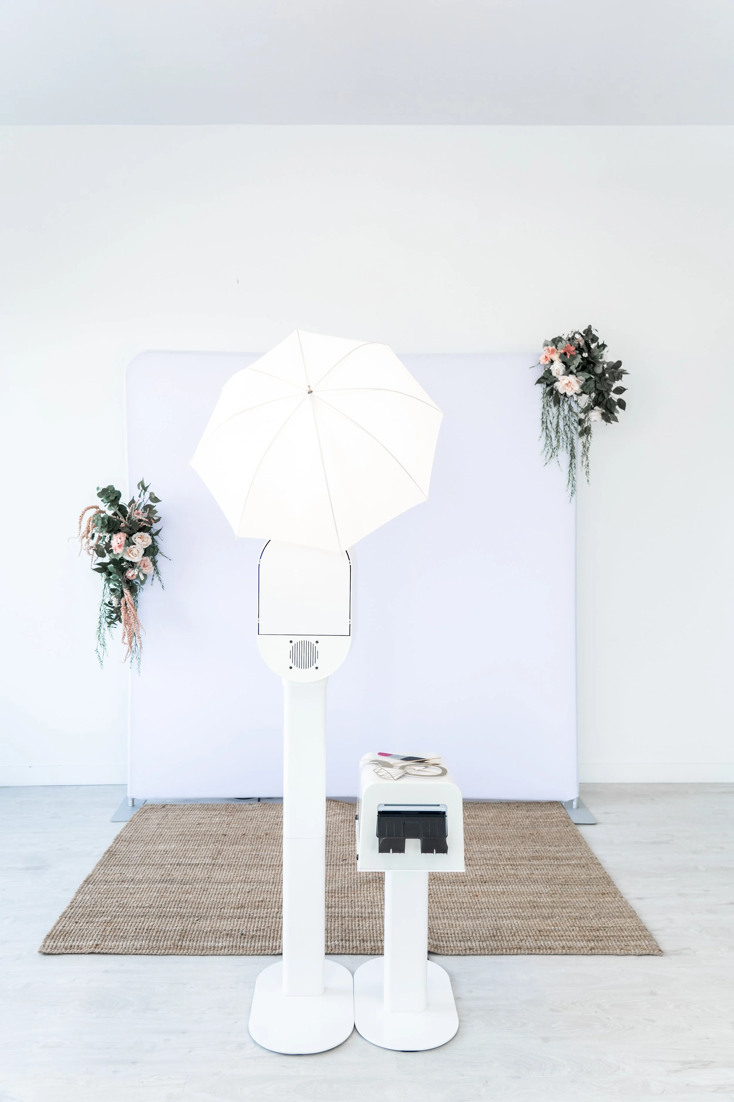 A white photo booth with a large white umbrella light, floral decorations on either side, a small white stand with a laptop and scissors, all set on a woven rug in a minimalistic room.
