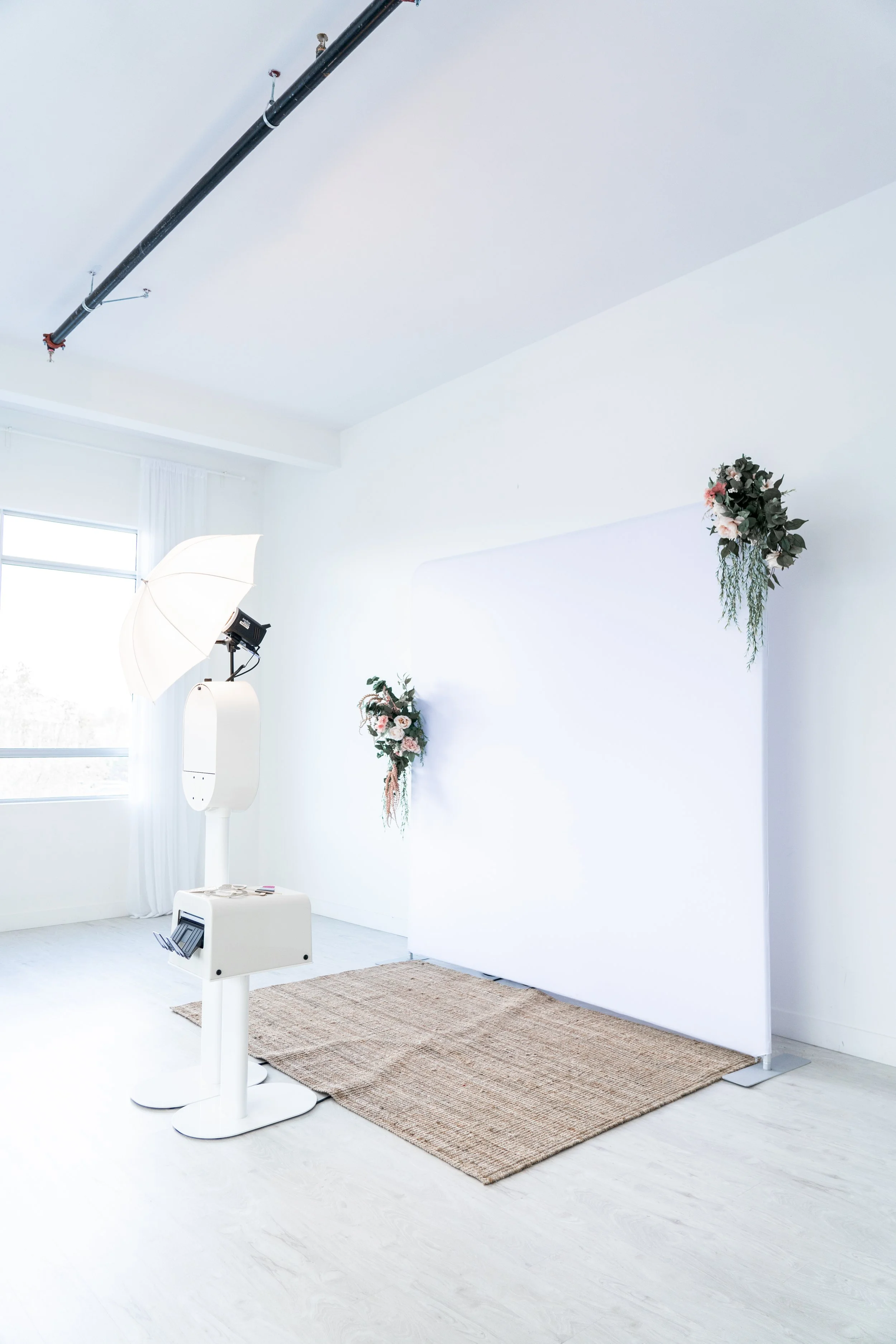 Photography studio setup with a white backdrop, floral decorations, soft box lighting, and a beige rug on white flooring.