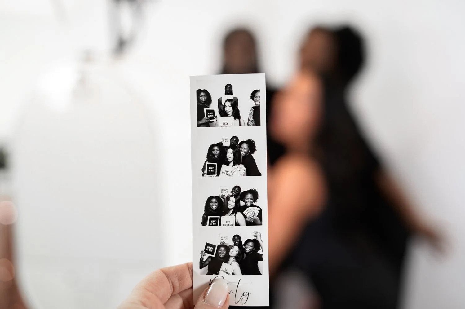 Black and white photo booth strip showing four images of three women having fun, taking photos, holding signs, and making playful expressions.
