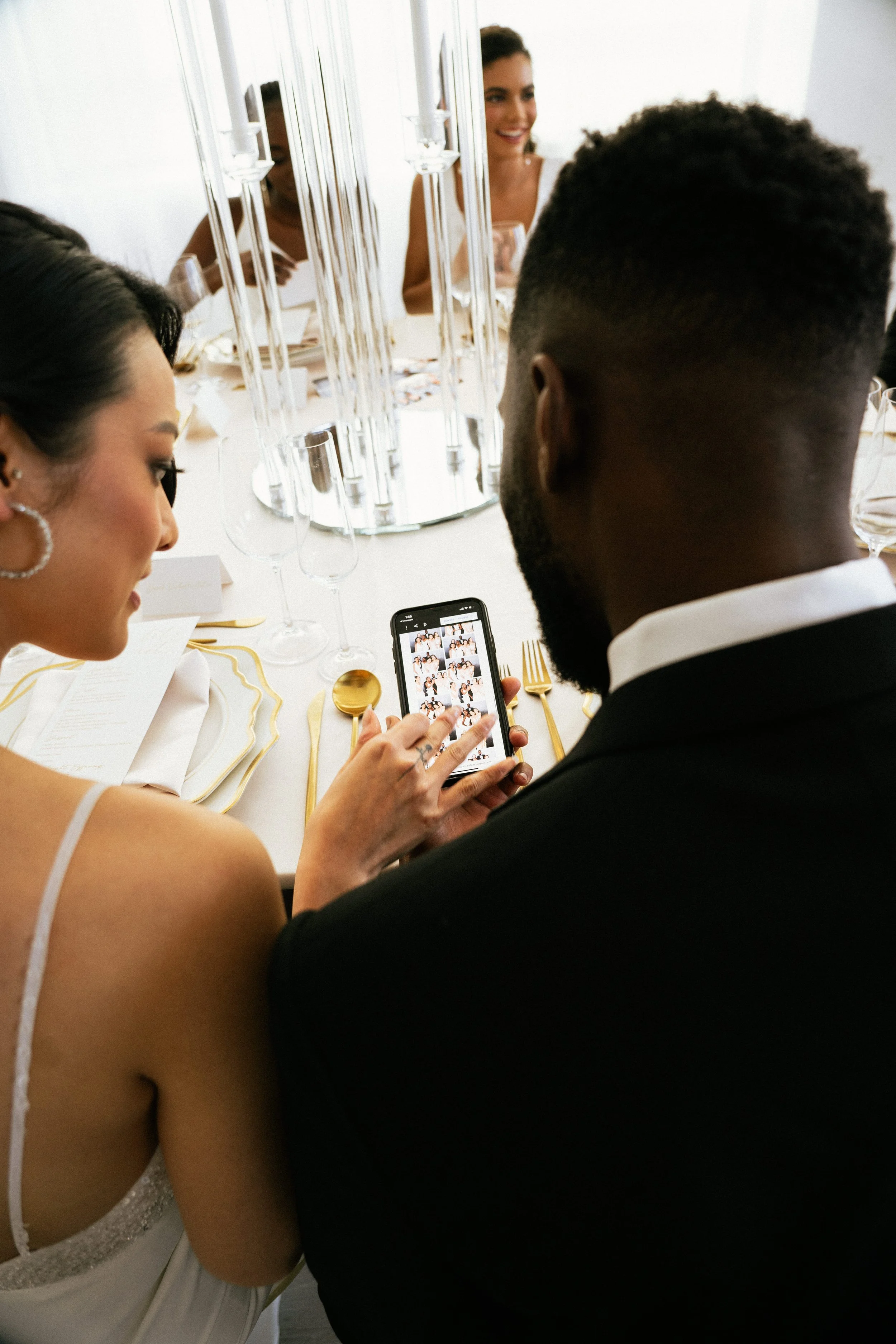 A woman in a white dress and a man in a black tuxedo sitting at a decorated table, taking a photo of themselves with a smartphone. In the background, other women are smiling.