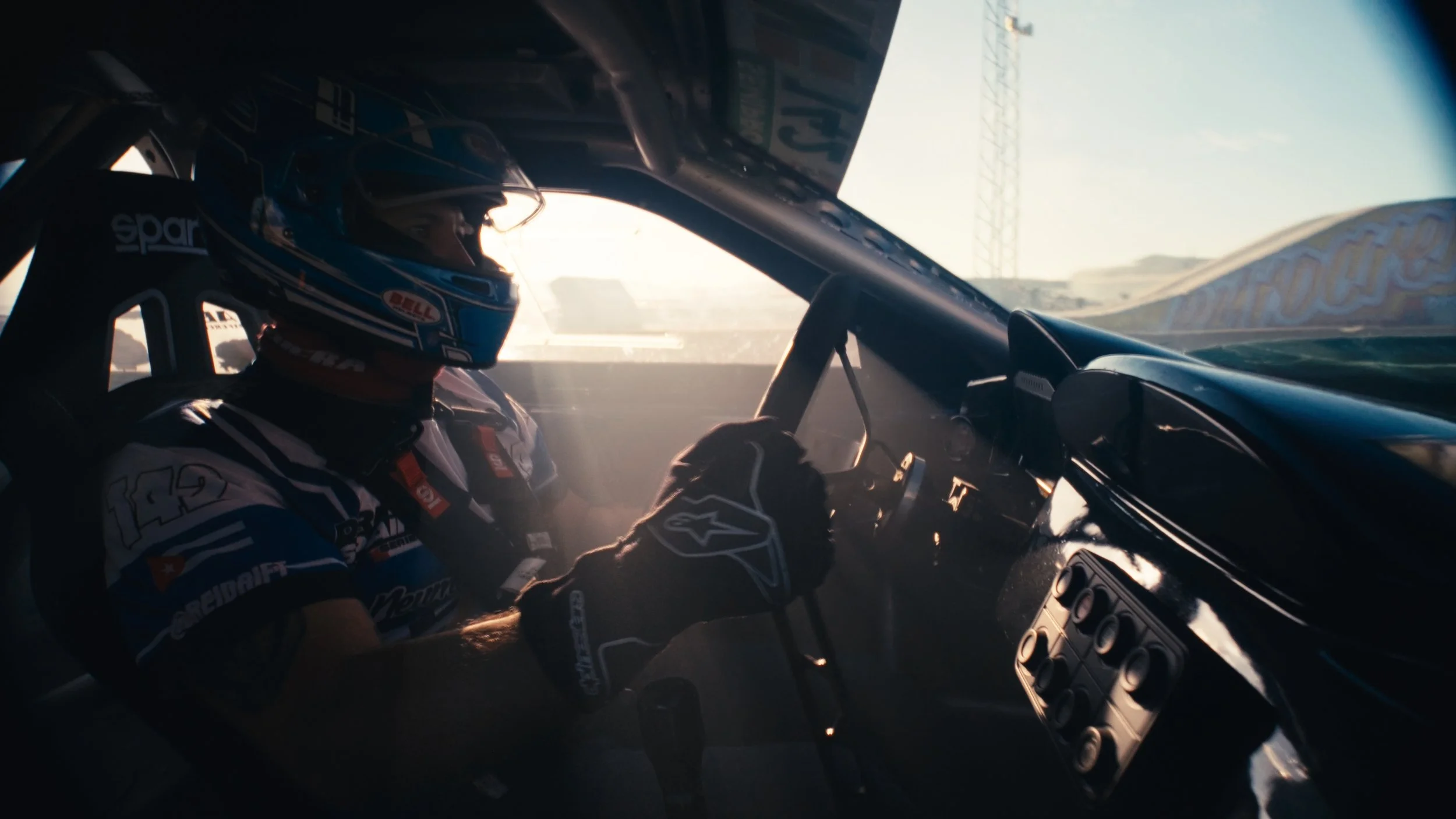 A race car driver wearing a blue helmet and racing suit sitting inside a race car.