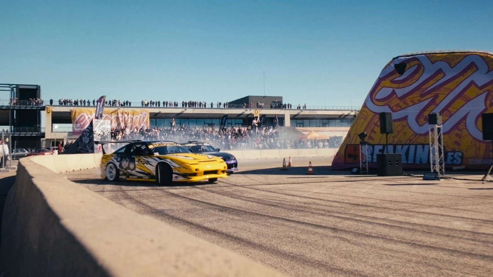 A yellow drift car with anime-style decals revolving around a corner on a racetrack, with spectators watching from the background and a large inflatable arch with promotional banners.