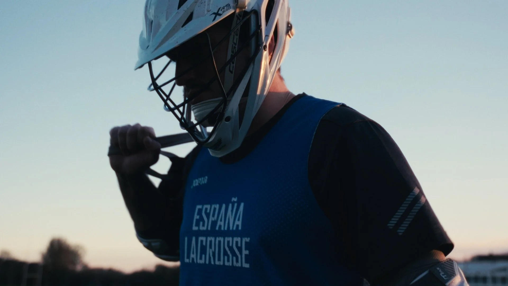 A lacrosse player wearing a white helmet and blue jersey with 'ESPAÑA LACROSSE' printed on it, holding a lacrosse stick set against a sunset sky.