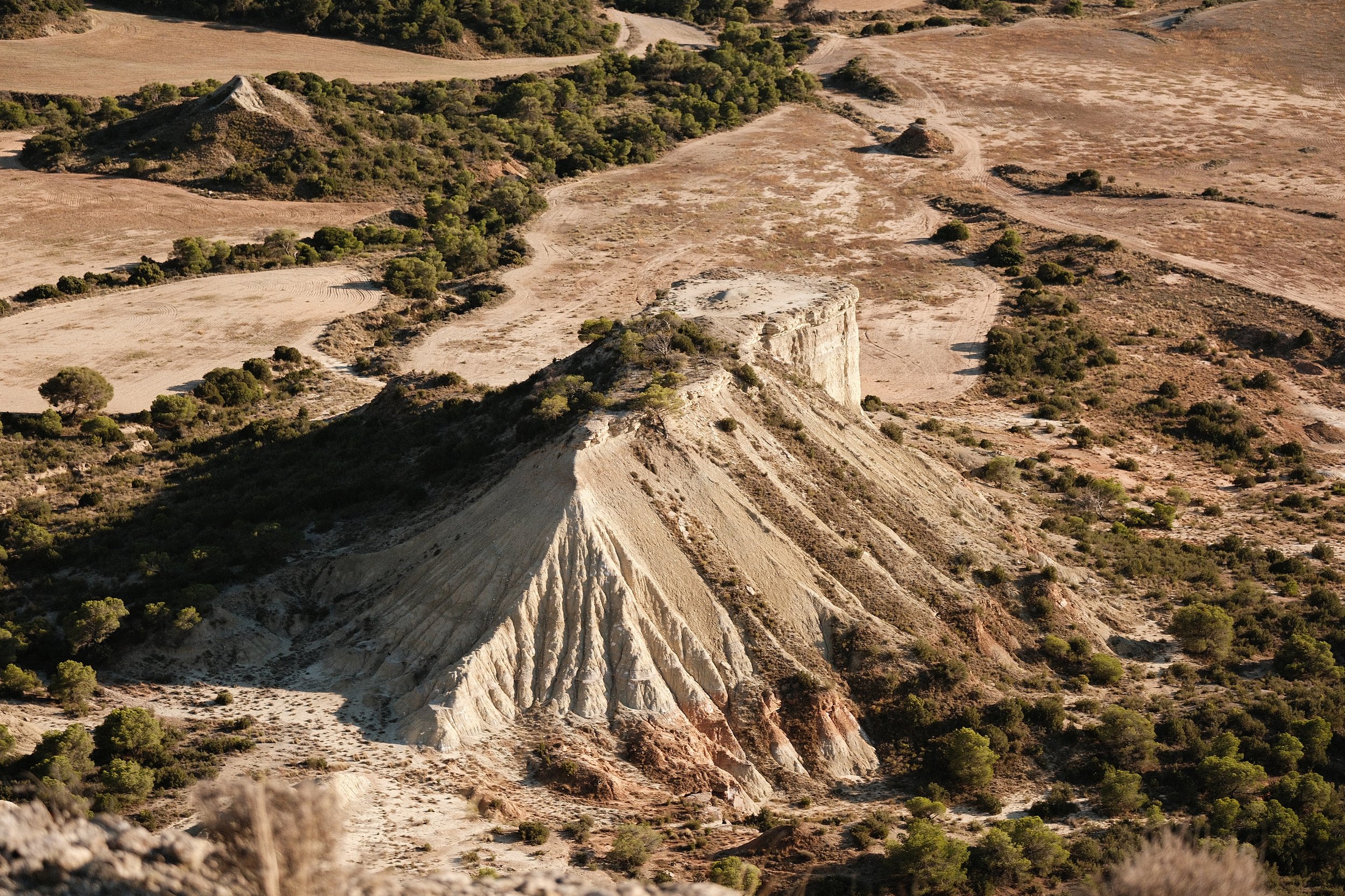 Aerial view of a dry, rugged landscape with a prominent white rock formation and some sparse green vegetation, including small trees and shrubs.