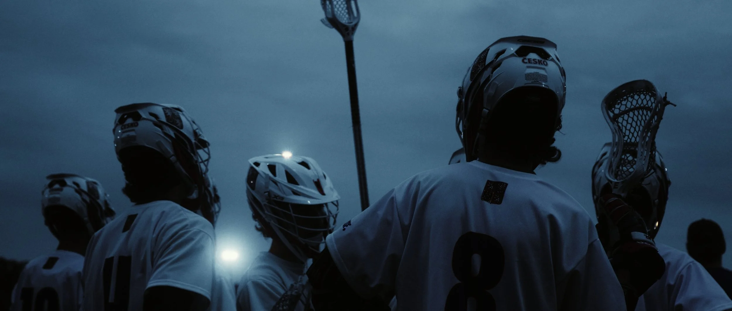 Lacrosse players in white uniforms and helmets standing together on the field during twilight or nighttime, with a dark sky and stadium lights in the background.
