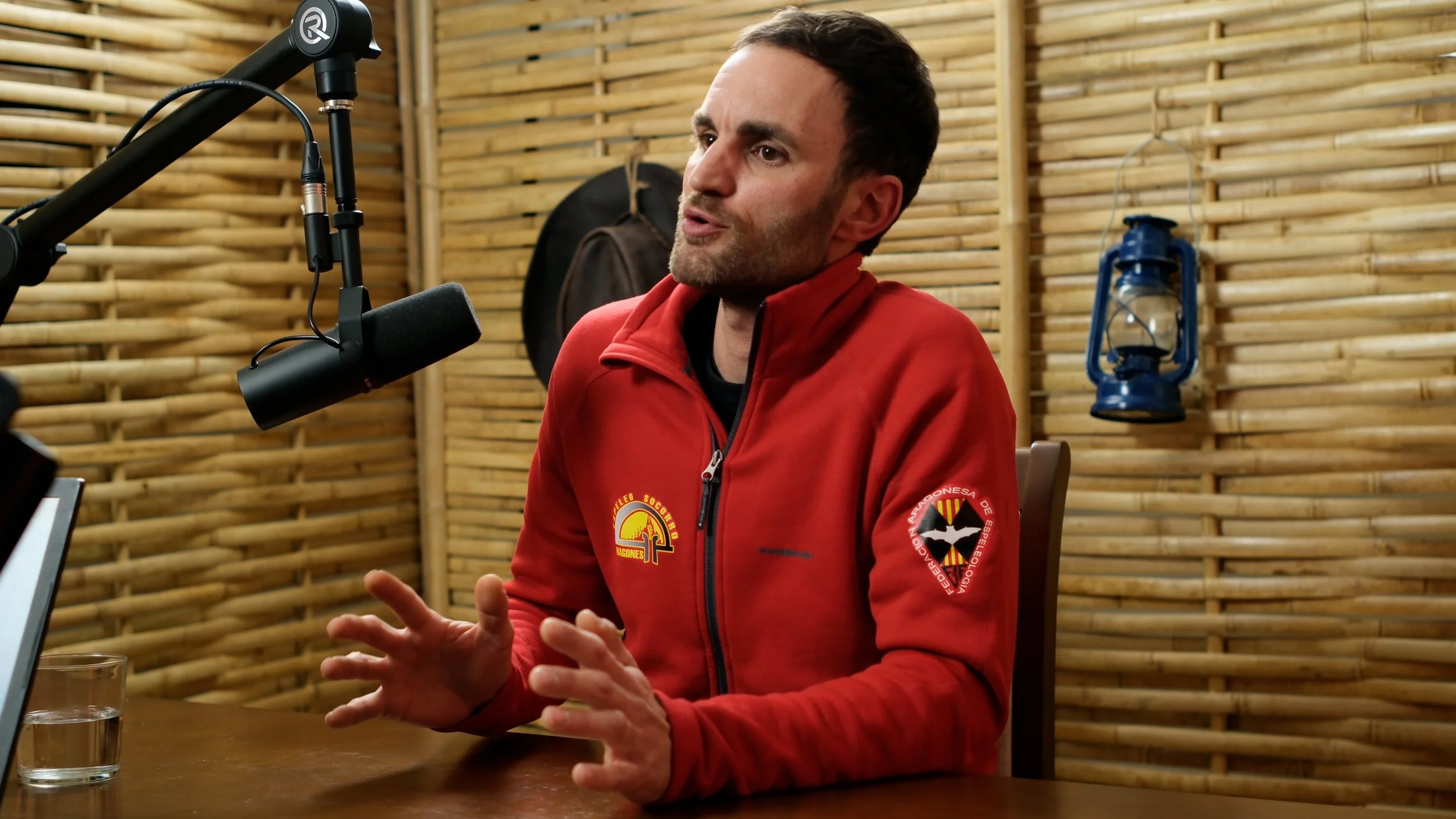 A man in a red jacket with logos on it is speaking into a microphone in a wooden-paneled room. There are glasses of water on the table in front of him, and a blue lantern is hanging on the wall behind him.