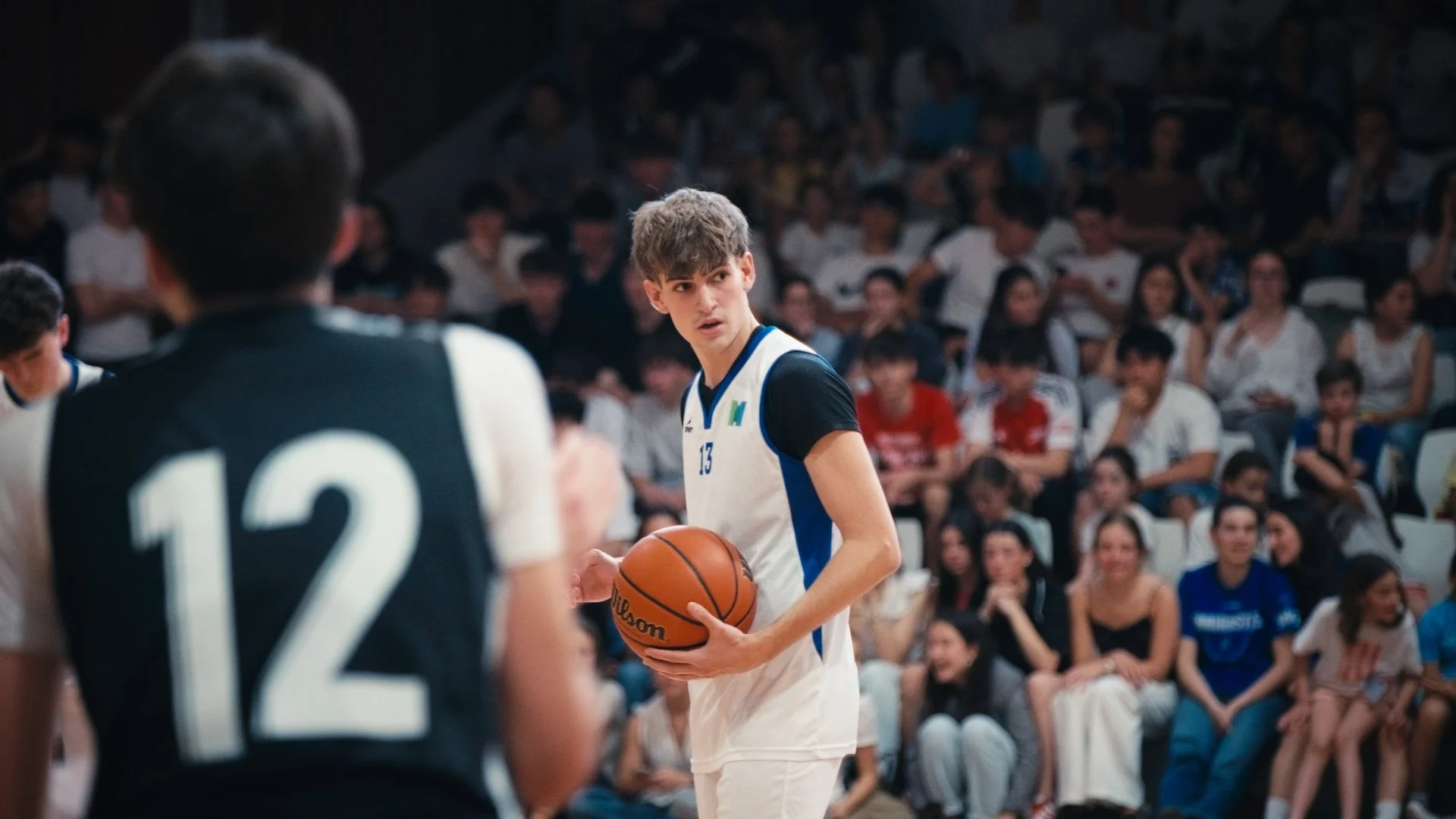 A young male basketball player wearing a white and blue jersey with the number 13, holding a basketball on a court during a game, with a crowd of spectators in the background.