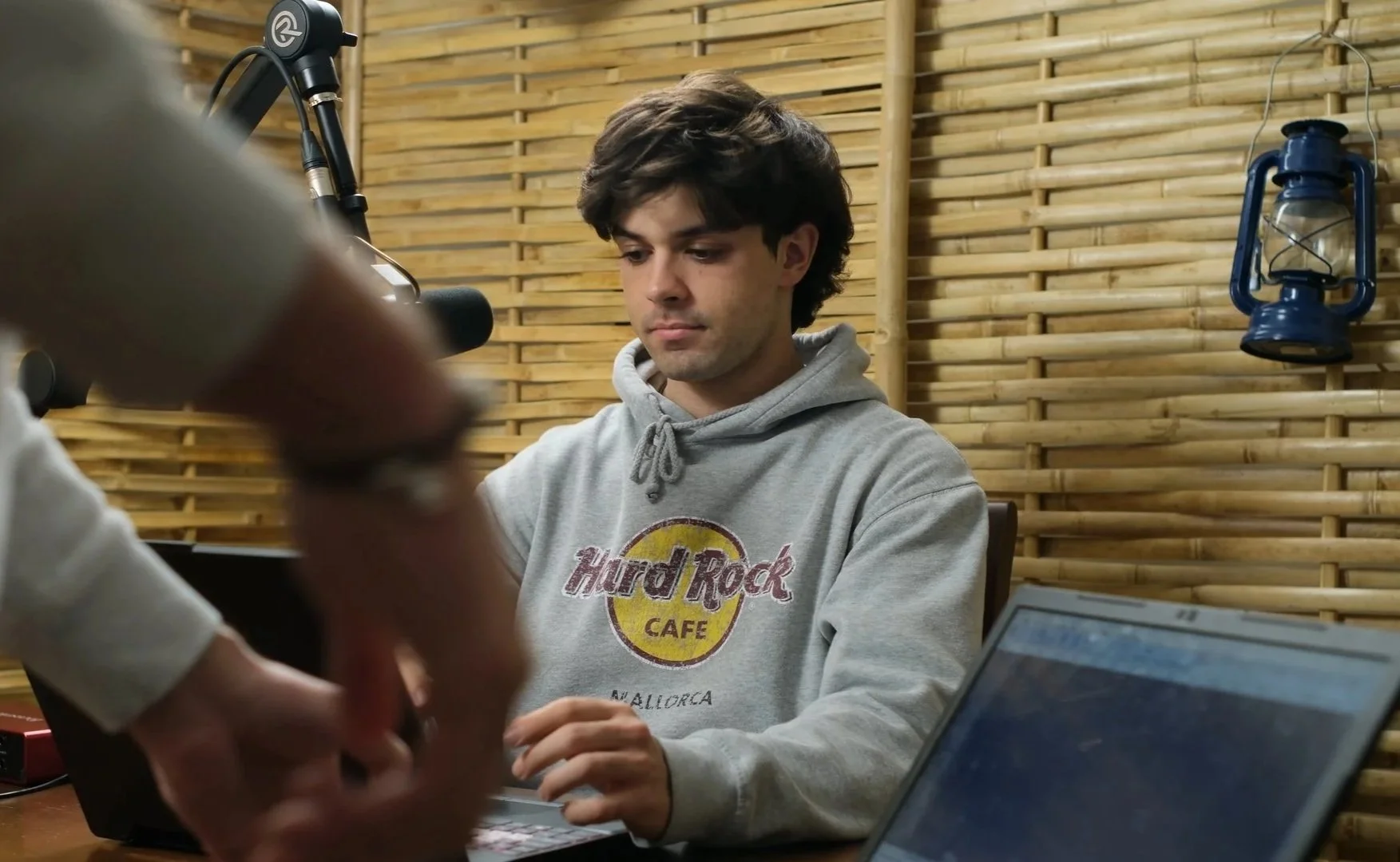 A young man in a gray hoodie with the Hard Rock Cafe logo sits at a desk with a laptop, in a room with a wooden slat wall and blue lanterns hanging on the wall.