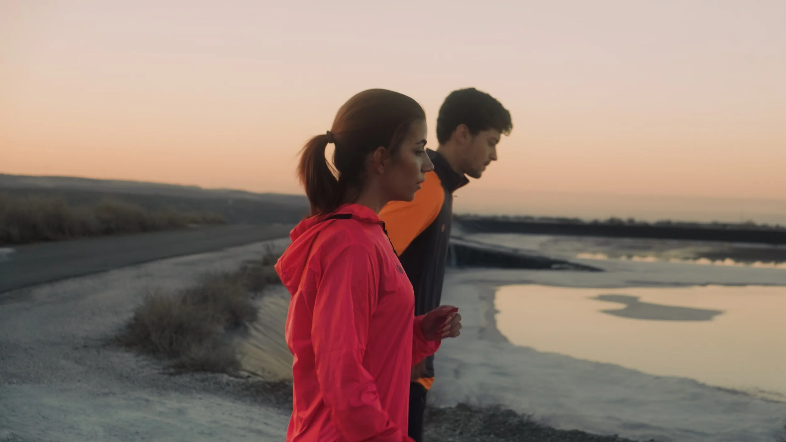 A woman in a pink jacket and a man in a dark jacket walk along a coastal area during sunset.
