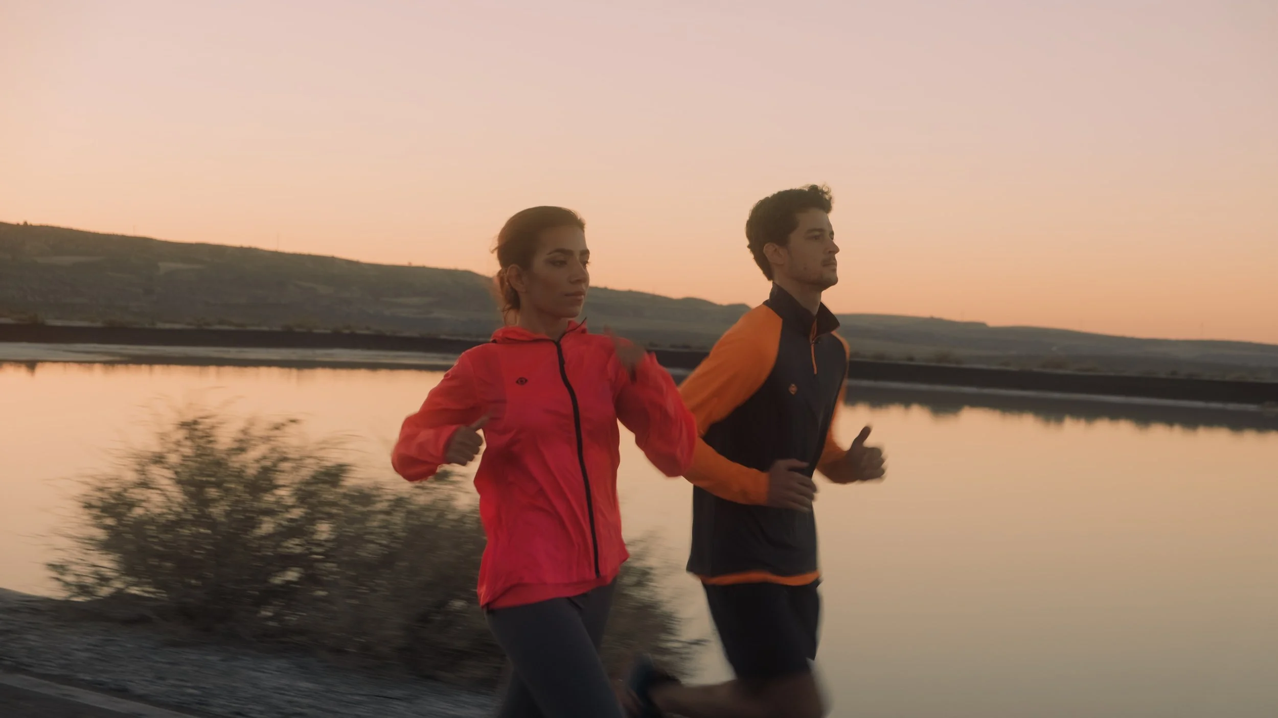 Two people running along a lakeside at sunset, with hills in the background.