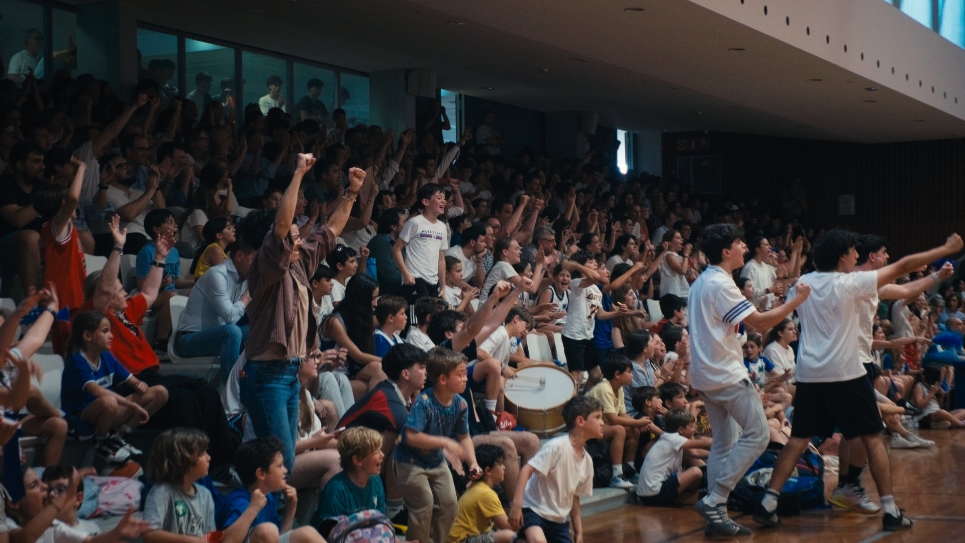 A large crowd of children and teenagers in an indoor auditorium, some standing, clapping, and dancing during a lively event.