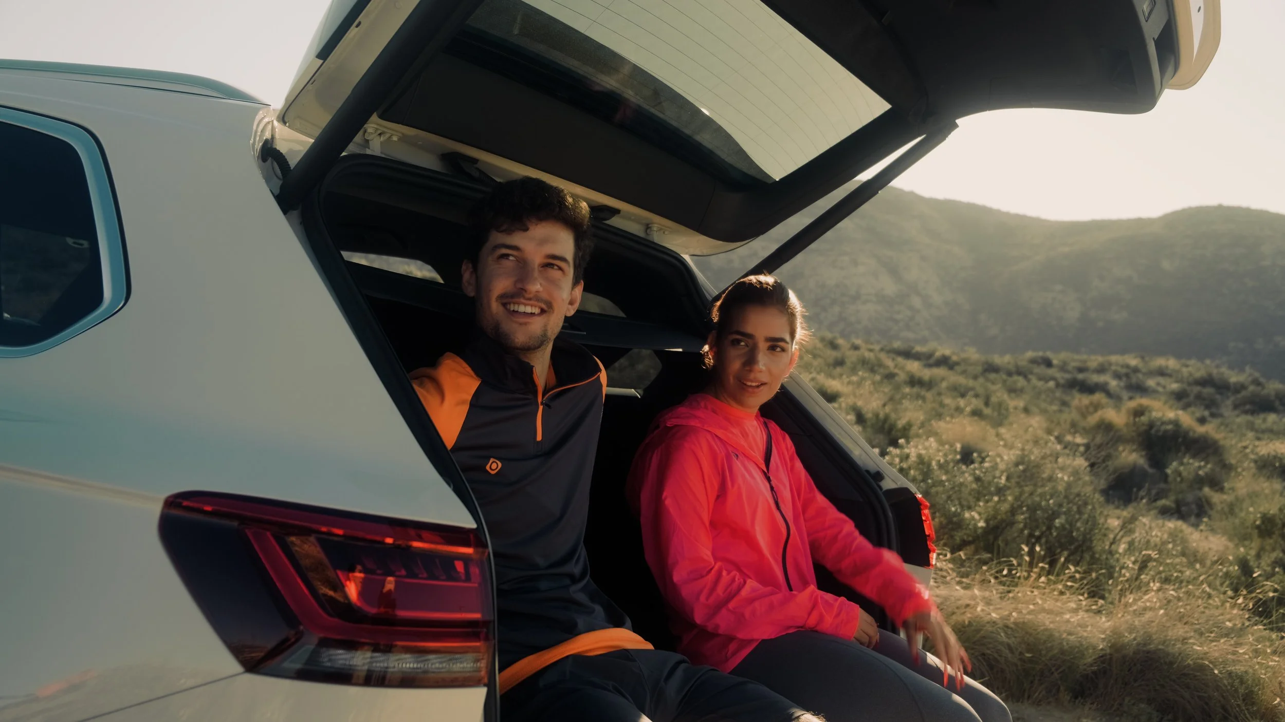 A man and woman sit in the back of a car with the hatch open, in a scenic outdoor setting with hills and greenery, during the daytime.