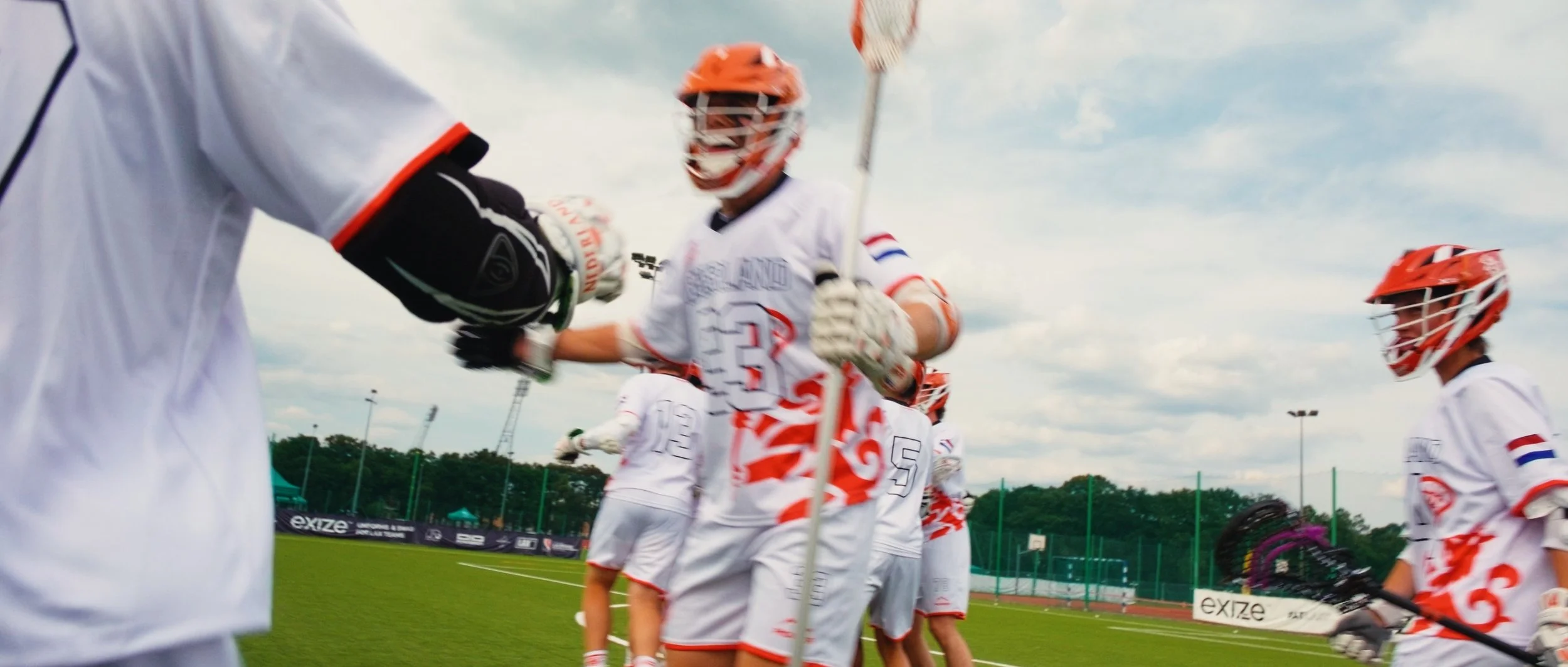Lacrosse players in white and red uniforms shaking hands on a field under a cloudy sky.