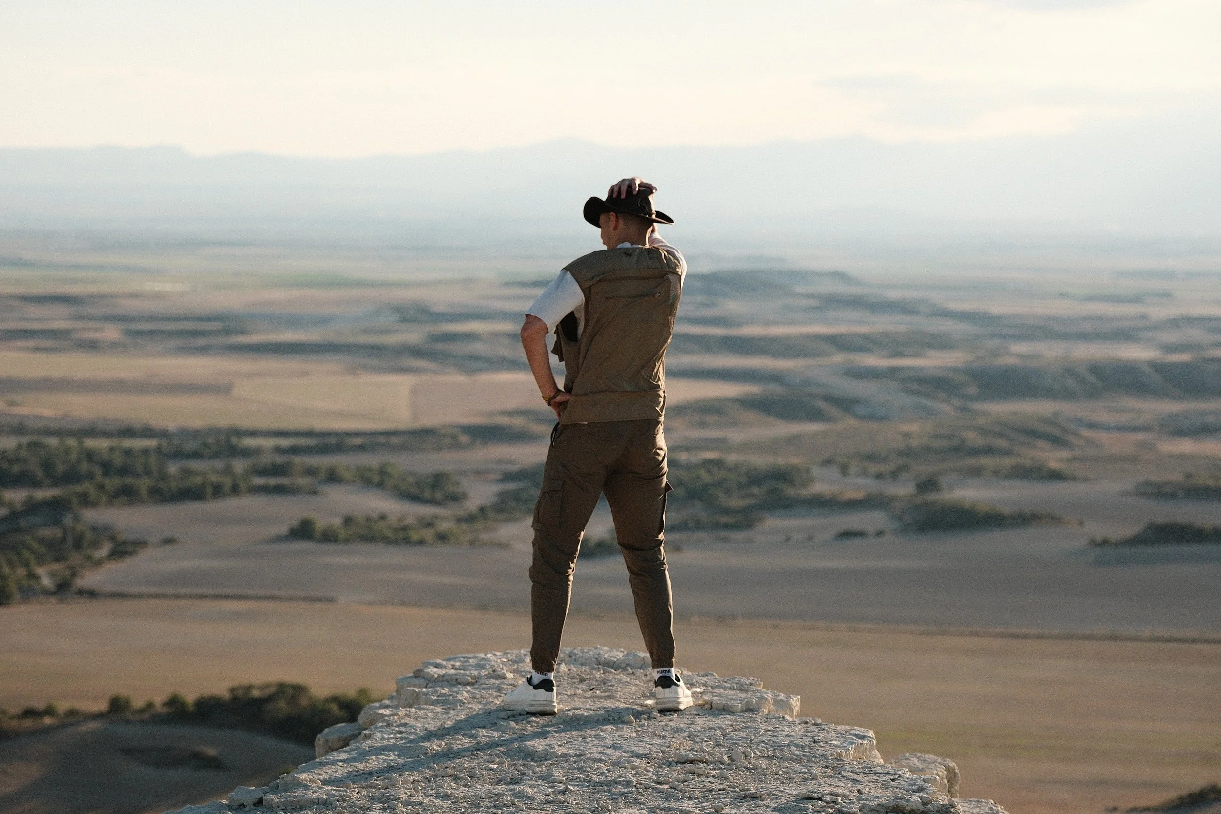 A person standing on a rocky ledge overlooking a vast landscape, wearing a cowboy hat, a vest, and white sneakers, with one hand on their head.