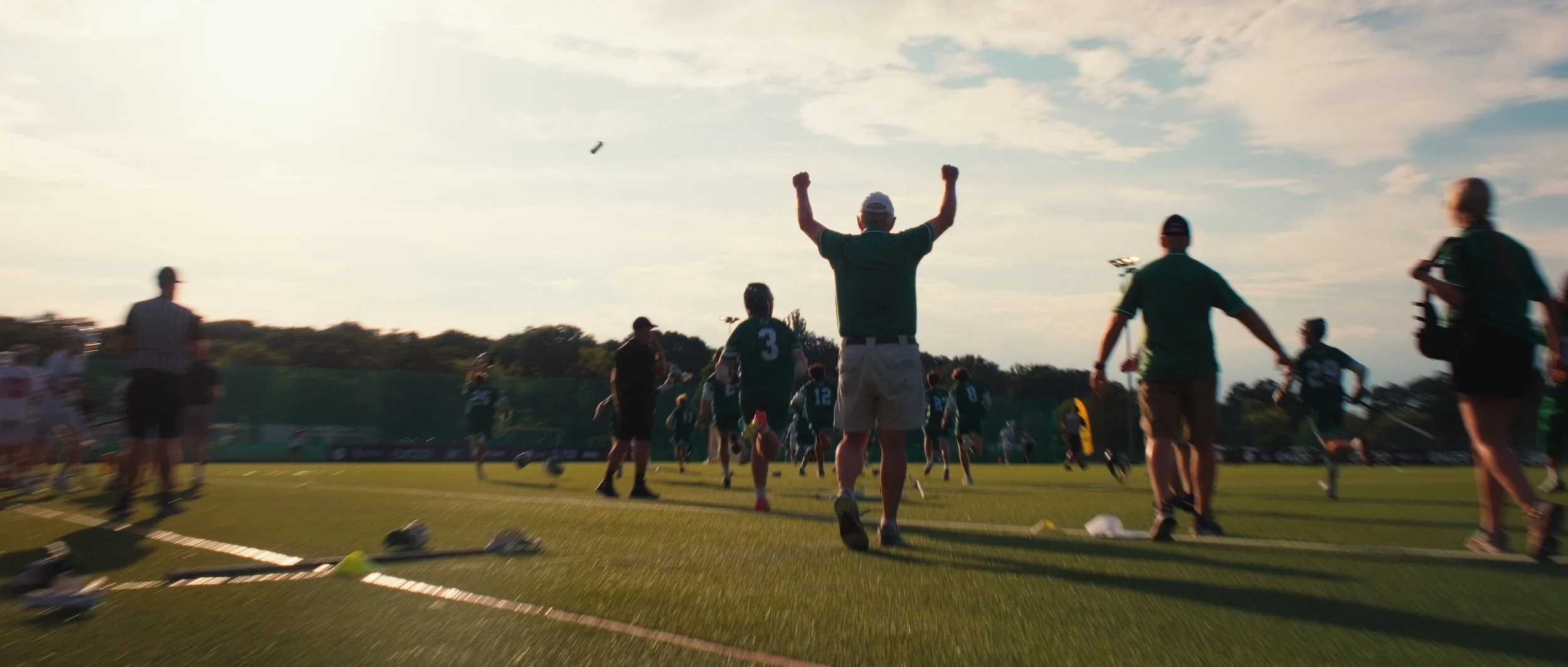 People celebrating on a sports field, with one person in the center raising their arms in victory, surrounded by others, under a partly cloudy sky.