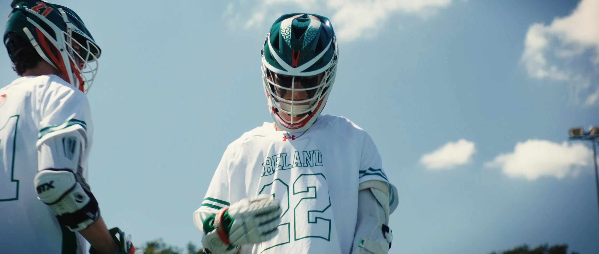 Lacrosse players wearing helmets and white jerseys with "Thailand" written on the front, standing outdoors under a partly cloudy sky.