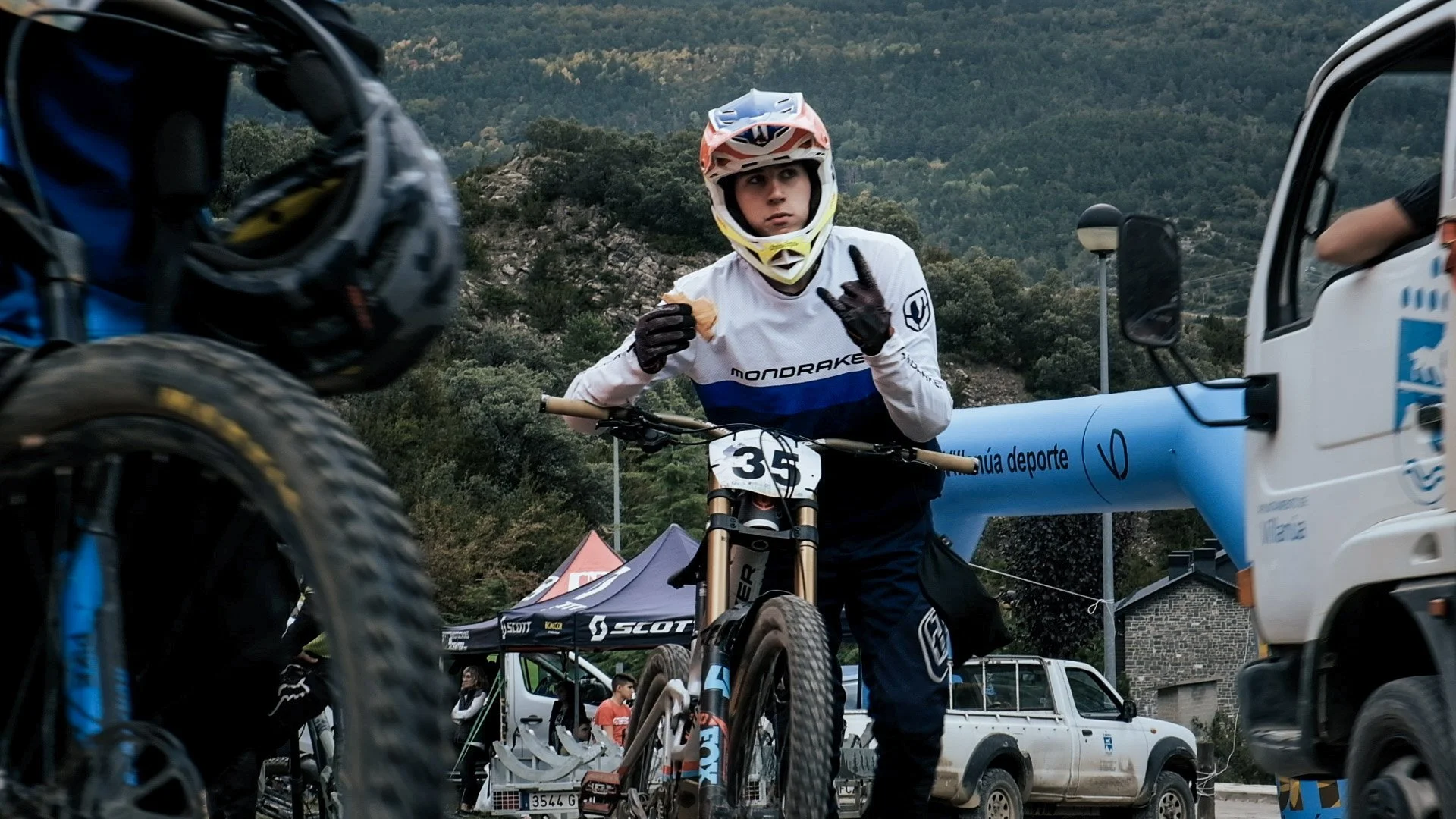 A mountain biker wearing a white jersey with blue and black accents, a helmet, and gloves stands holding his bike at an outdoor event. Race number 35 is attached to the front of his bike. There are vehicles, tents, and people in the background with t