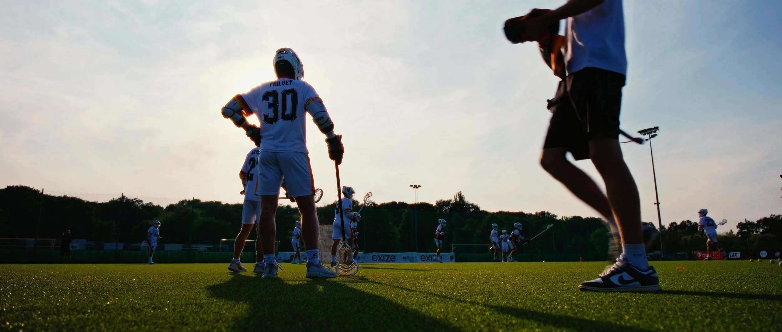 Lacrosse players practicing on a field during sunset, some holding lacrosse sticks, wearing helmets and uniforms.