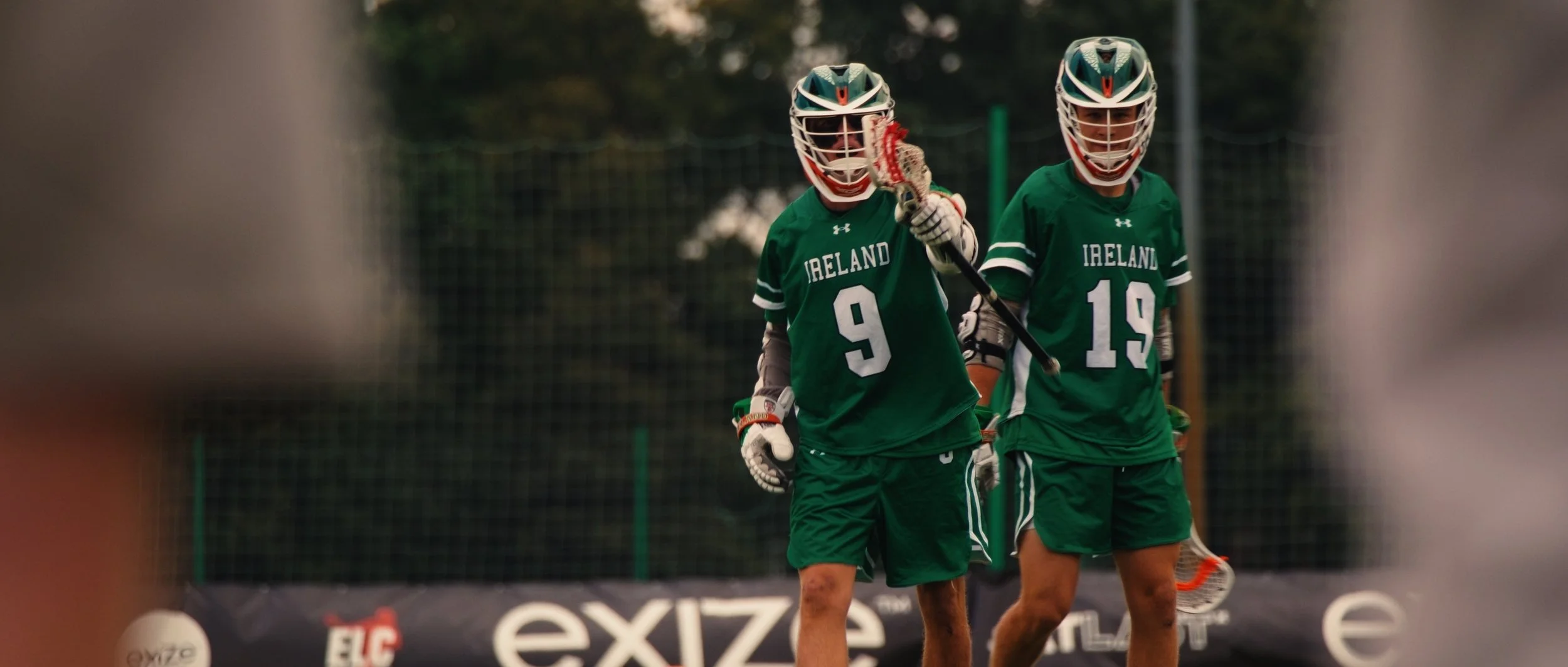 Two Irish men in green sports uniforms and helmets, holding lacrosse sticks on a field, with goal netting and trees in the background.
