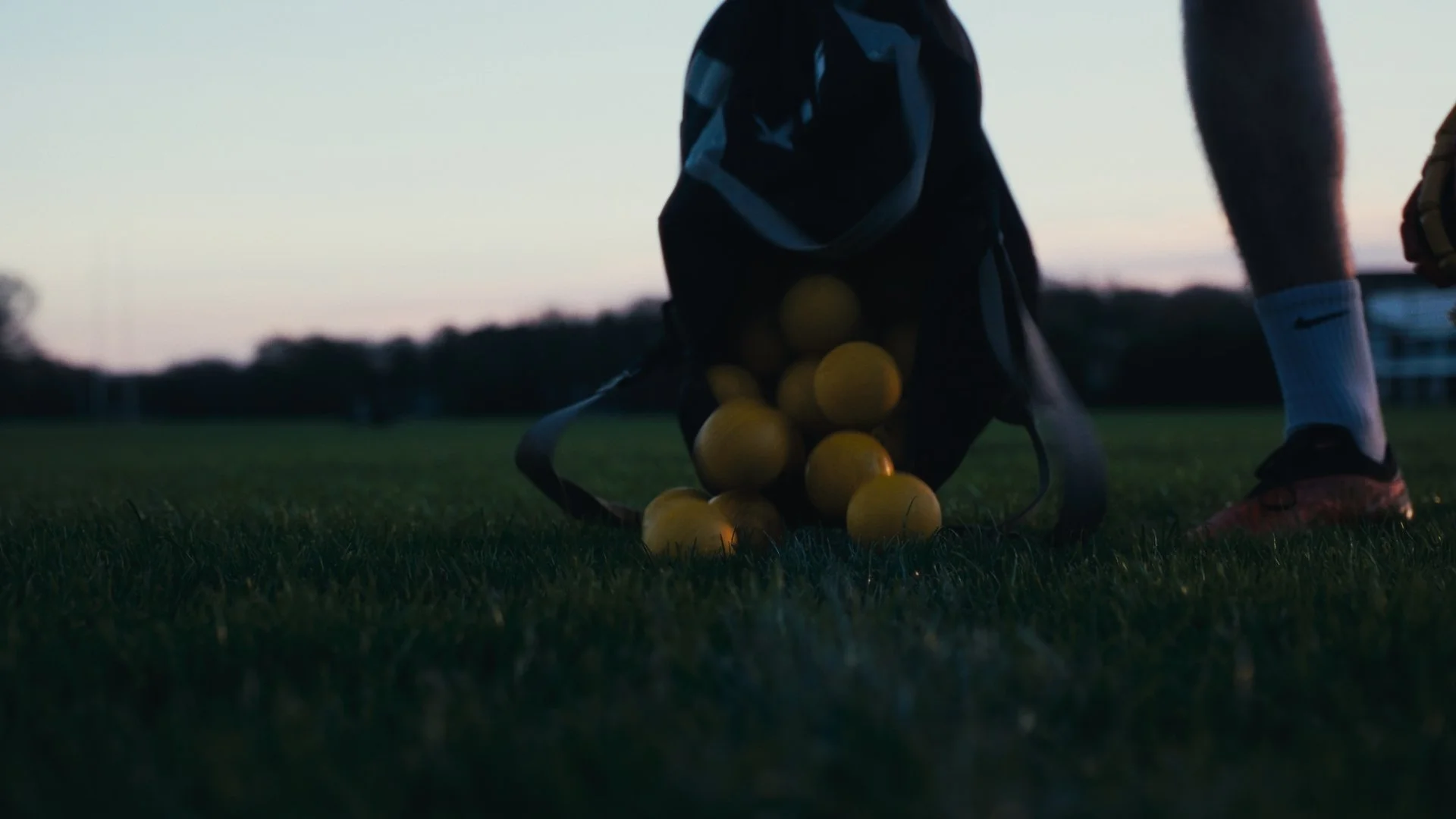 A black sports backpack on a grassy field with yellow tennis balls spilling out, with a person's legs wearing socks and orange cleats nearby, at dusk or dawn.