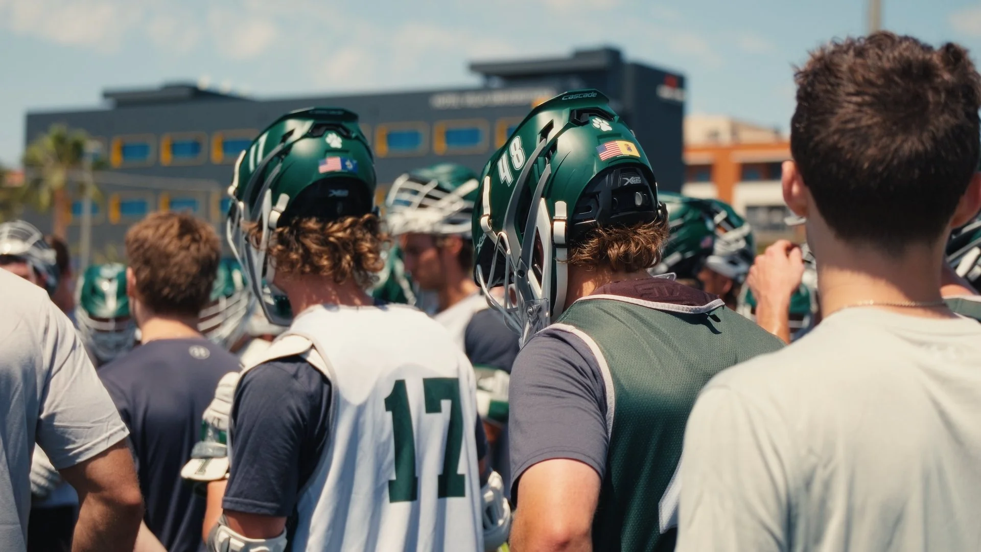 Young football players wearing green helmets and uniforms are gathered outdoors, possibly during a game or practice, with a building and palm trees in the background on a sunny day.
