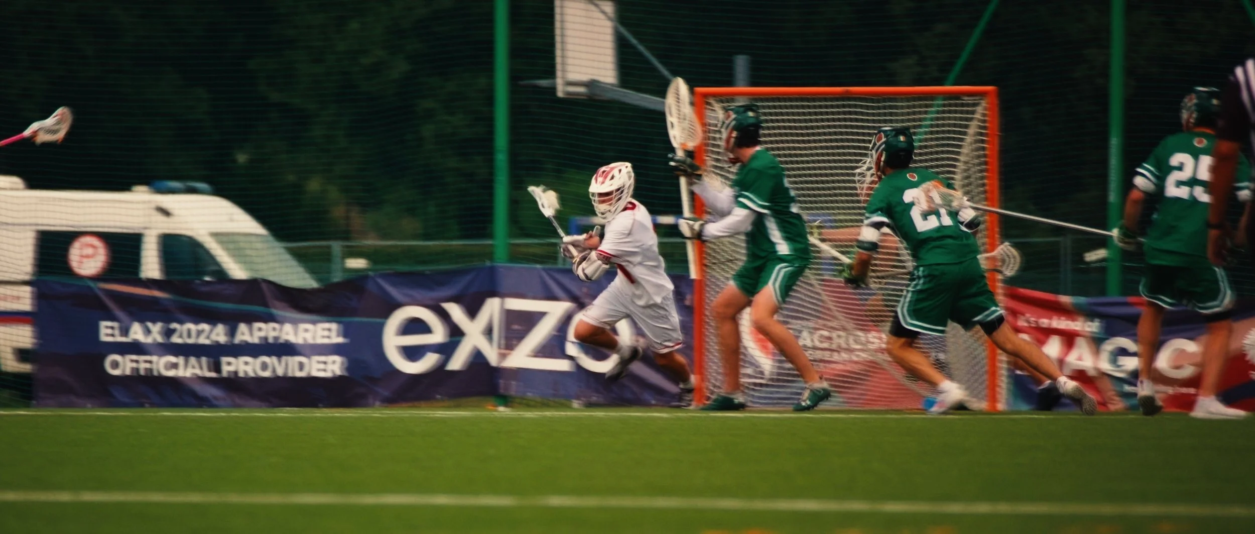 Lacrosse players in green and white uniforms playing near goal; one player in white with red accents handling lacrosse stick near goal, while two players in green attempt to block or defend.