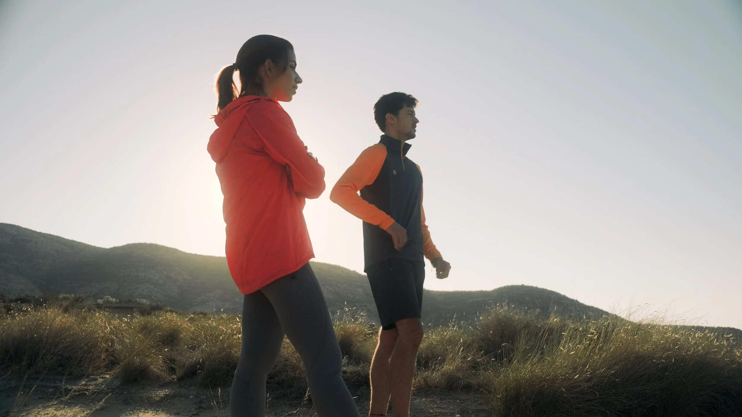 Two people hiking outdoors with hills in the background, one woman wearing a red jacket and a man in a black and orange jacket, both walking on grassy terrain during late afternoon or early evening.