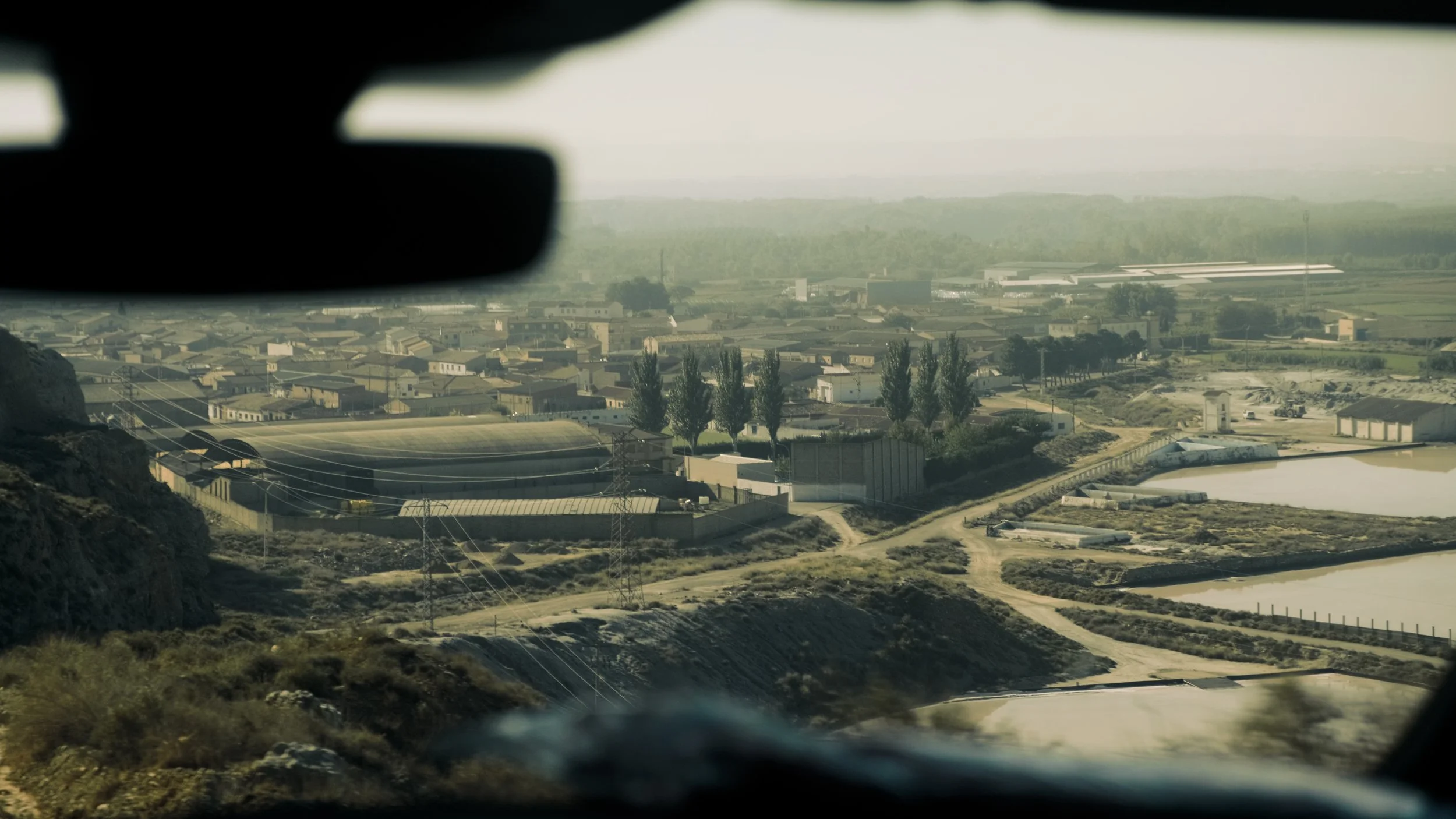 A view of a town or industrial area seen from inside a vehicle, with some plants and water bodies in the foreground and hazy hills in the background.