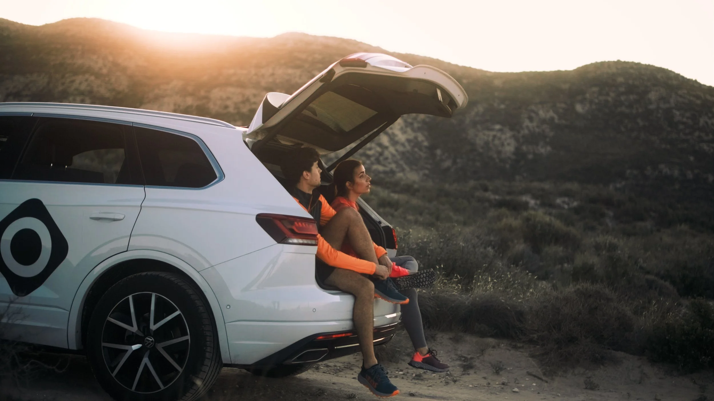 A man and woman sitting in the open trunk of a white SUV, parked in a desert landscape at sunset.