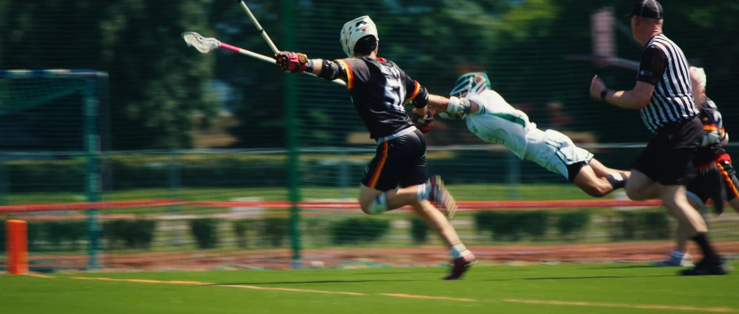 Lacrosse players in action on the field during a game, with one player diving to avoid being hit by the ball while another player reaches out with a stick.