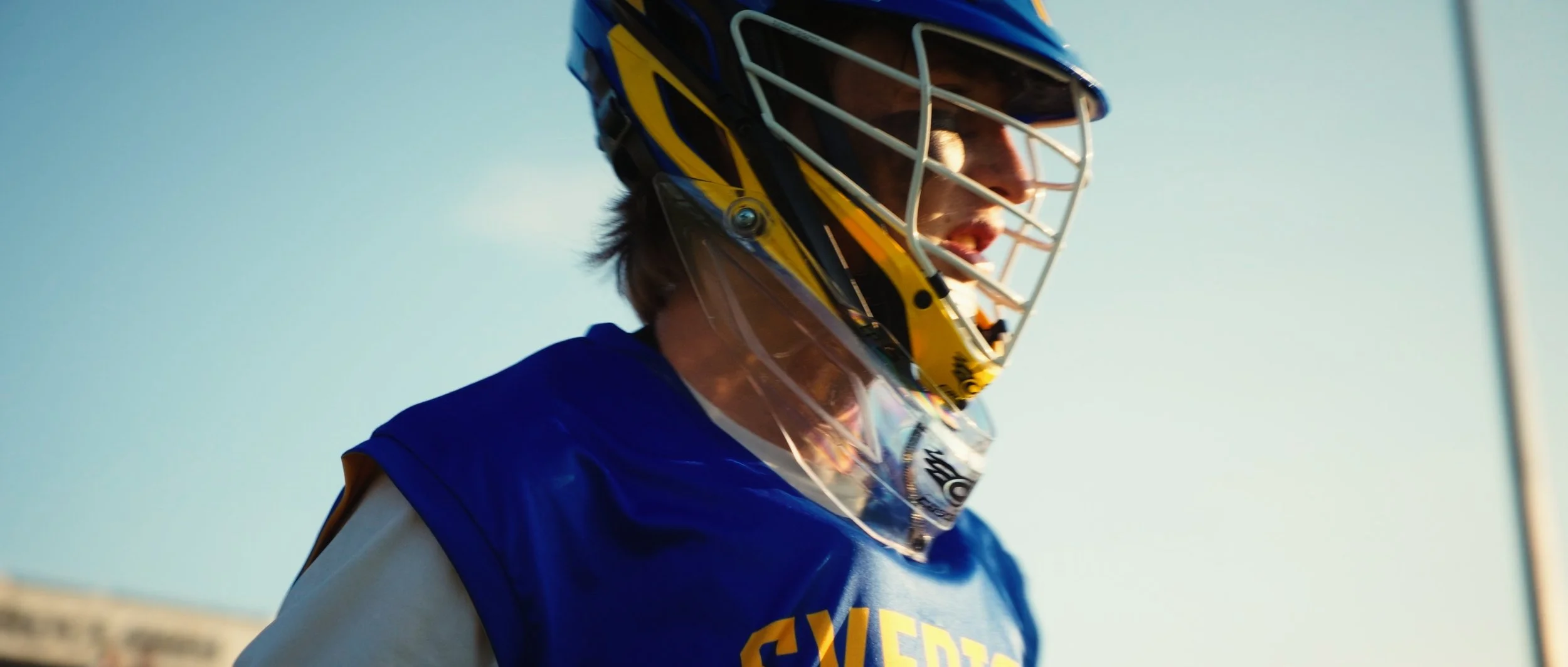 A lacrosse goalie wearing a helmet and protective gear, looking to the right against a clear sky.