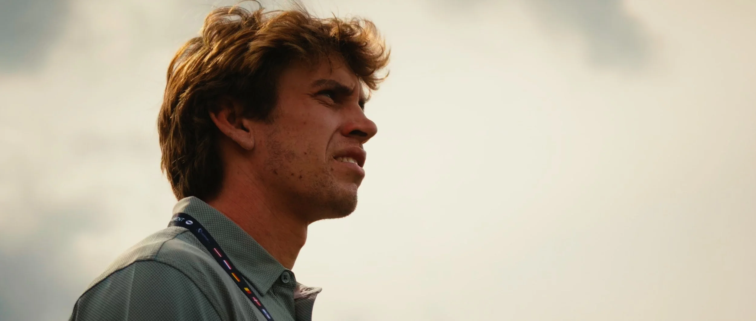 Side profile of a young man with tousled brown hair, wearing a collared shirt and a lanyard, looking into the distance against a cloudy sky.