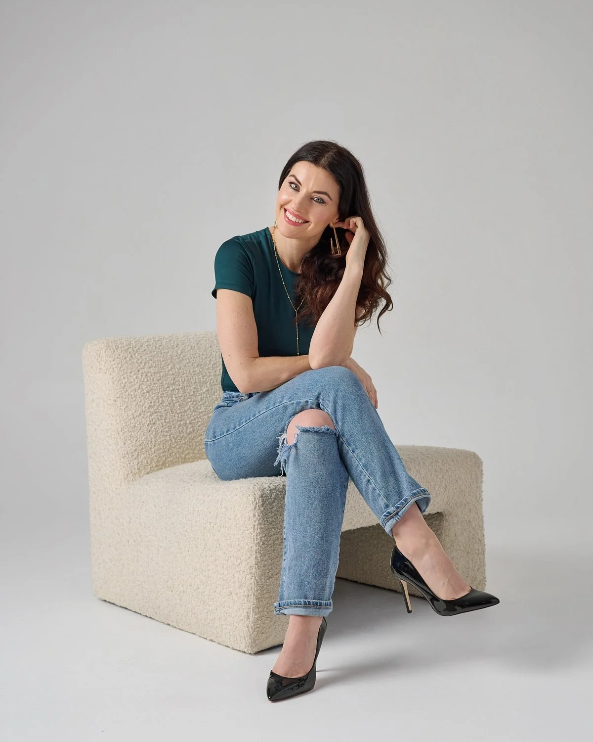 Stylish female therapist with long brown hair wearing ripped jeans, golden jewelry, and black heels, while seated in a plush beige chair.