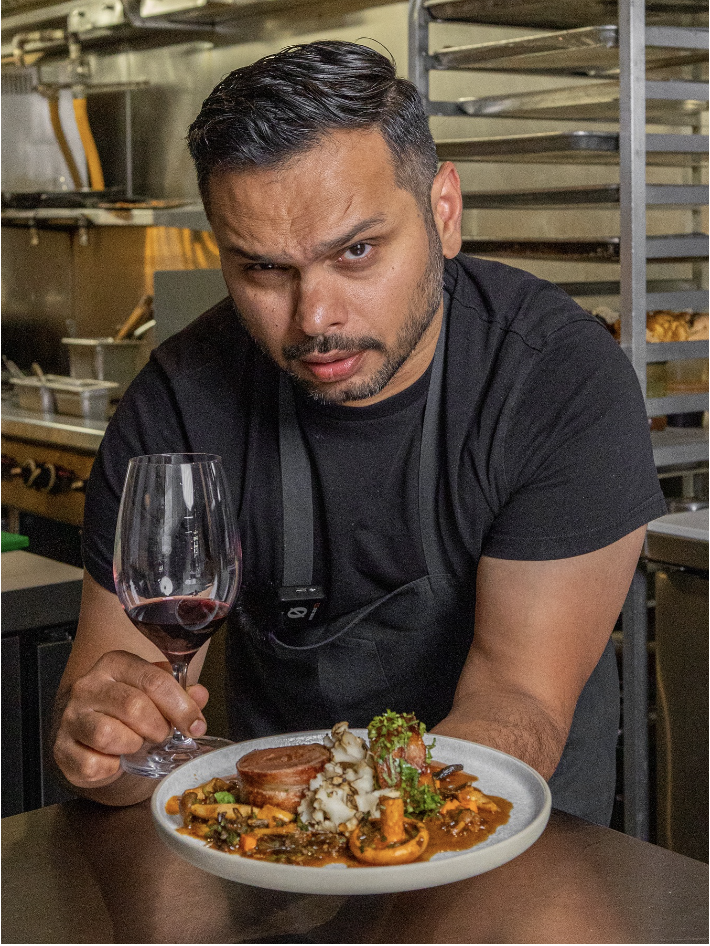 Chef Miheer is holding a glass of red wine and presenting his Signature Dish in a restaurant kitchen.