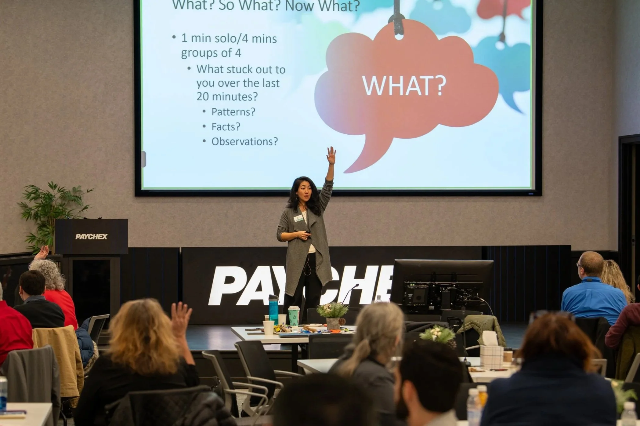 Woman standing on stage presenting to group with a slide in the background saying What?