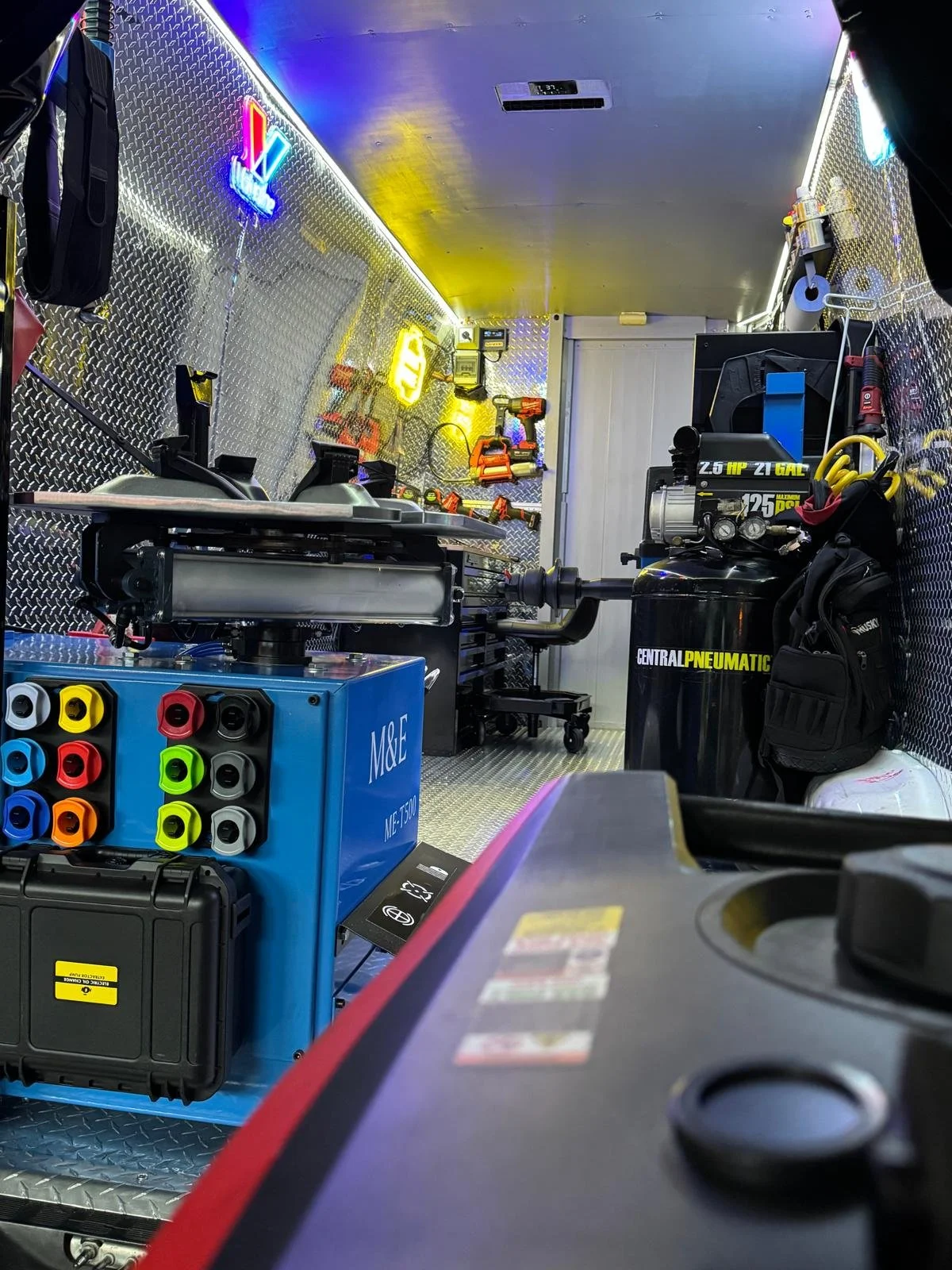 Inside of a service or repair truck with tools, equipment, and supplies neatly arranged, including colored connectors, drills, a pneumatic compressor, and disorganized tools on the diamond plate walls and ceiling.