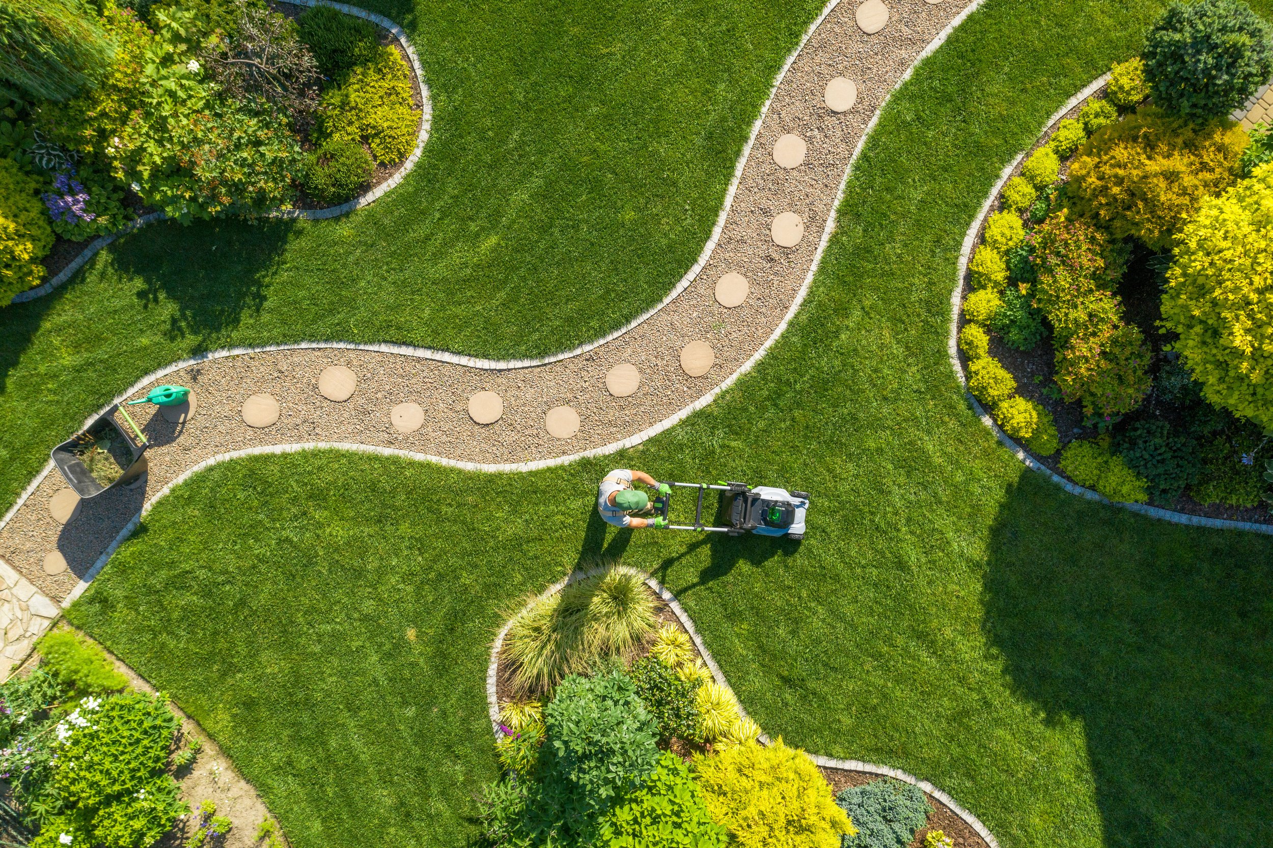 A person mowing a green lawn in a garden with a pathway, bushes, trees, and a garden bench.