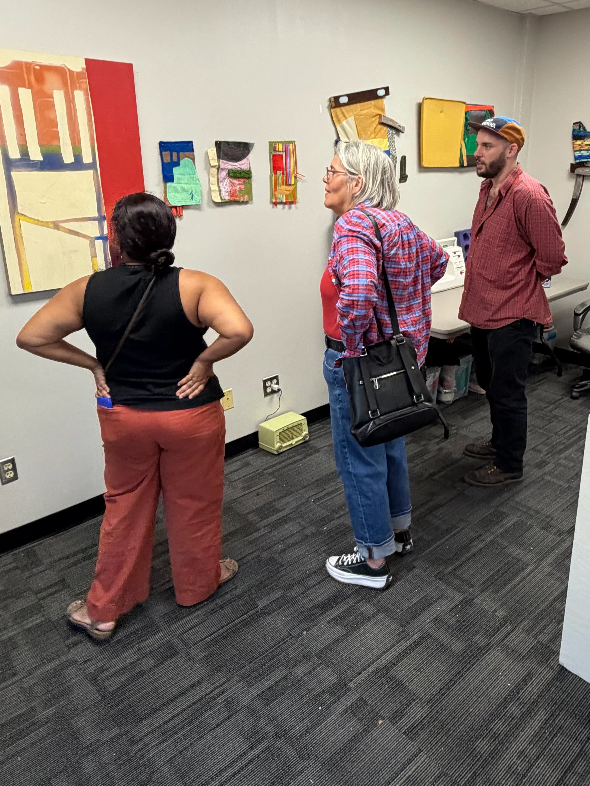 Joshua Bryant stands in front of a wall of his artwork as two visitors speak to him