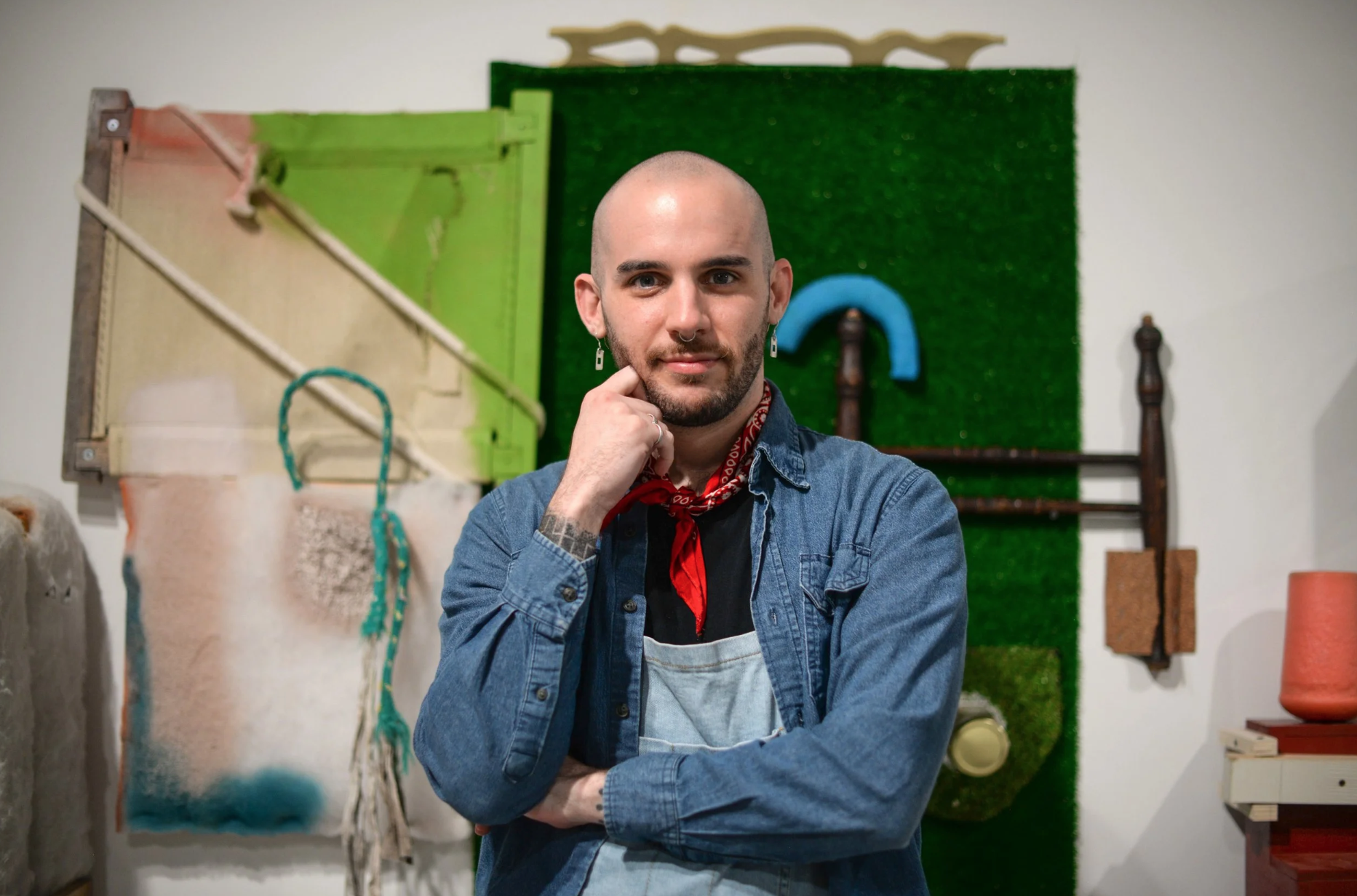 A non-binary person wearing a denim button down shirt and a red bandana looks thoughtfully at the camera