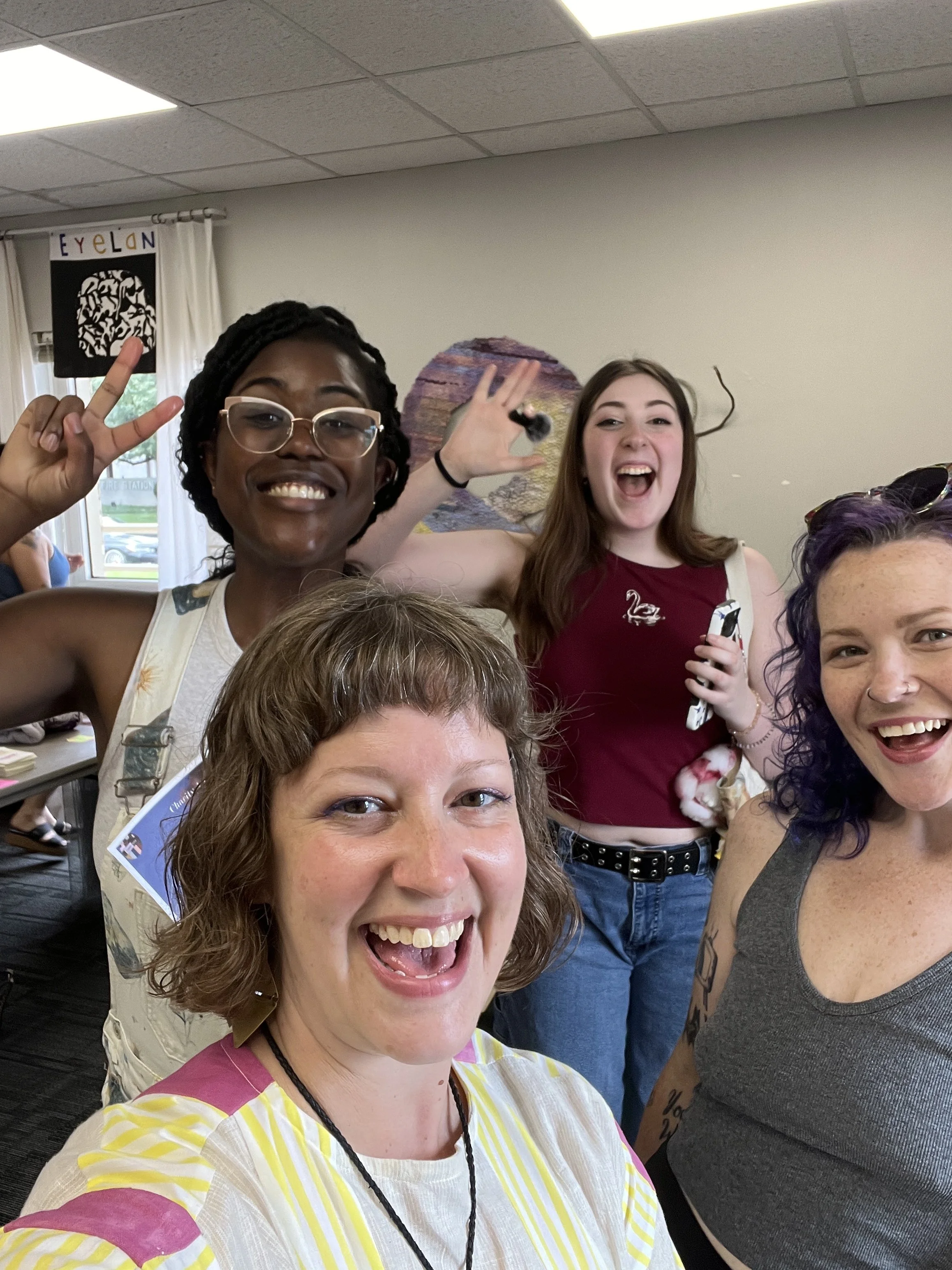 A white woman with brown wavy hair takes a selfie in an art studio with three young artists smile in the background