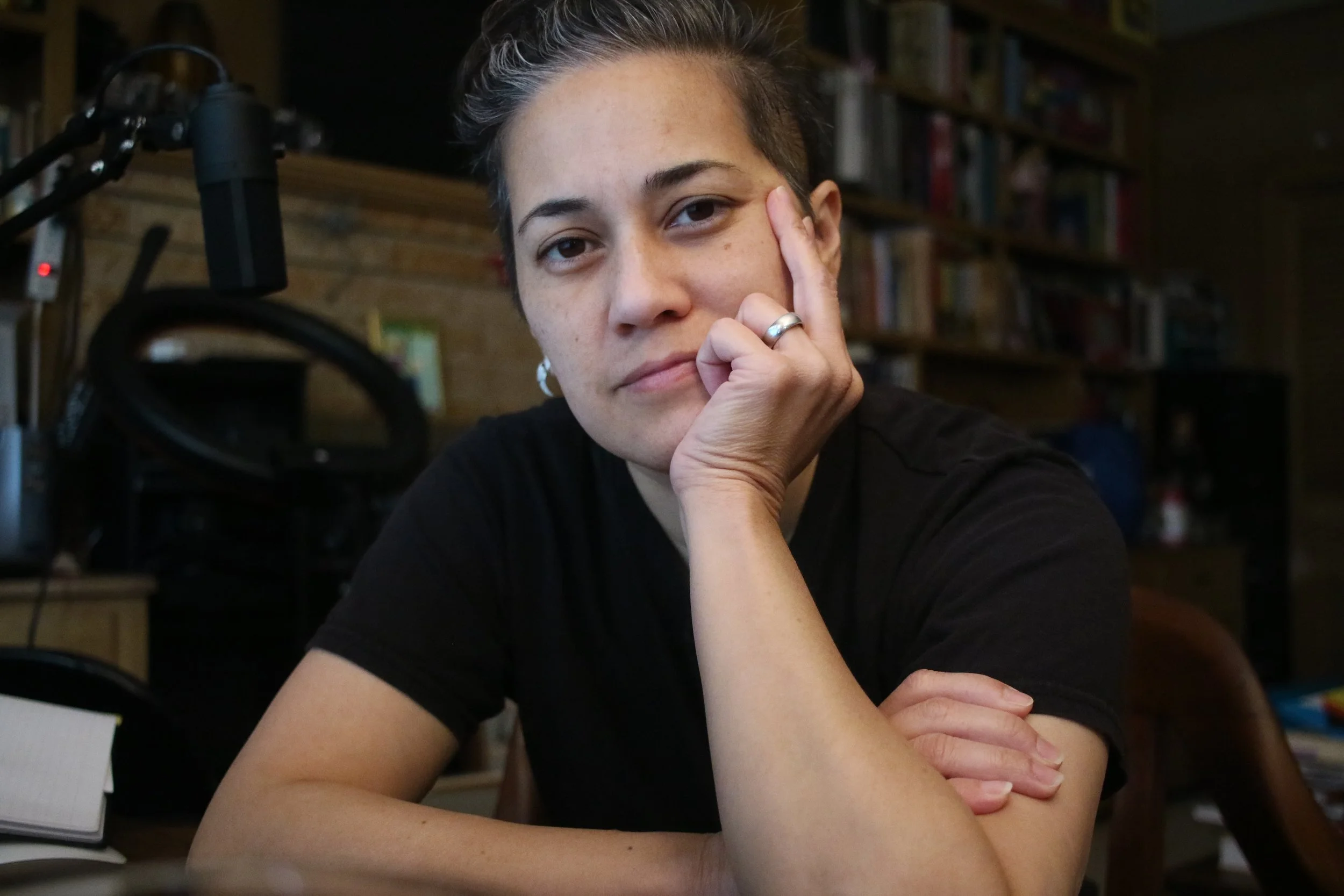 A person with olive skin wearing a black tshirt sits at a desk and looks thoughtfully at the camera
